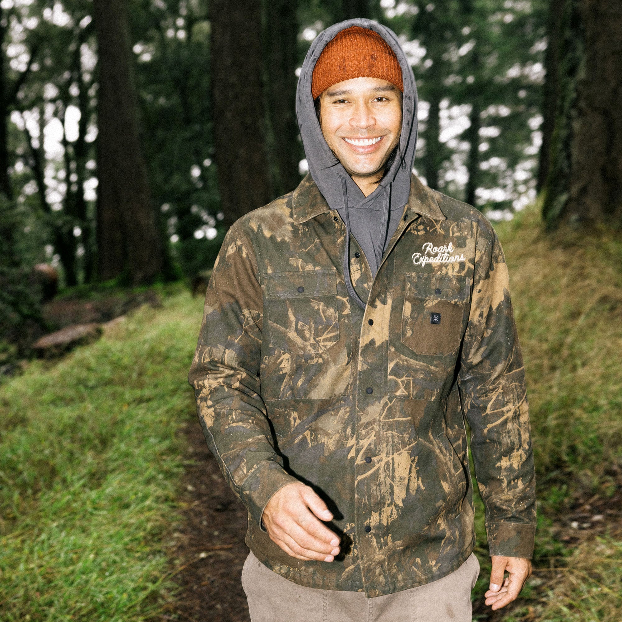 Man wearing a camouflaged Hebrides unlined jacket with hood, designed for winter adventures, blending into a natural outdoor setting.