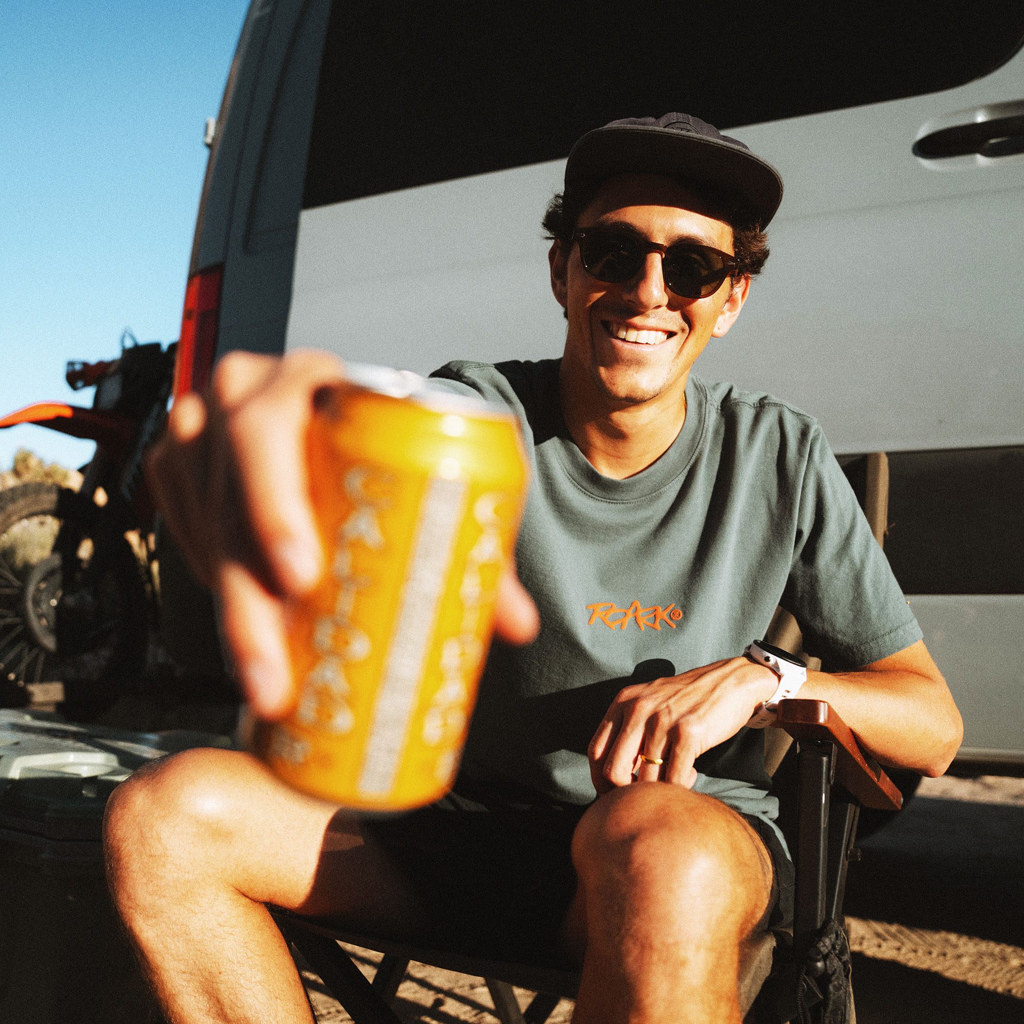 Man wearing sage green Kimo tee with sunglasses and black cap holds yellow beverage can outdoors near white van