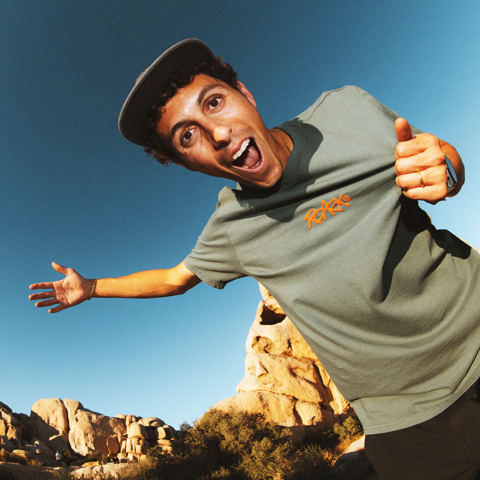Man wearing slate green Roark Homegrown Premium Tee with orange logo, posing outdoors against blue sky and rocky landscape