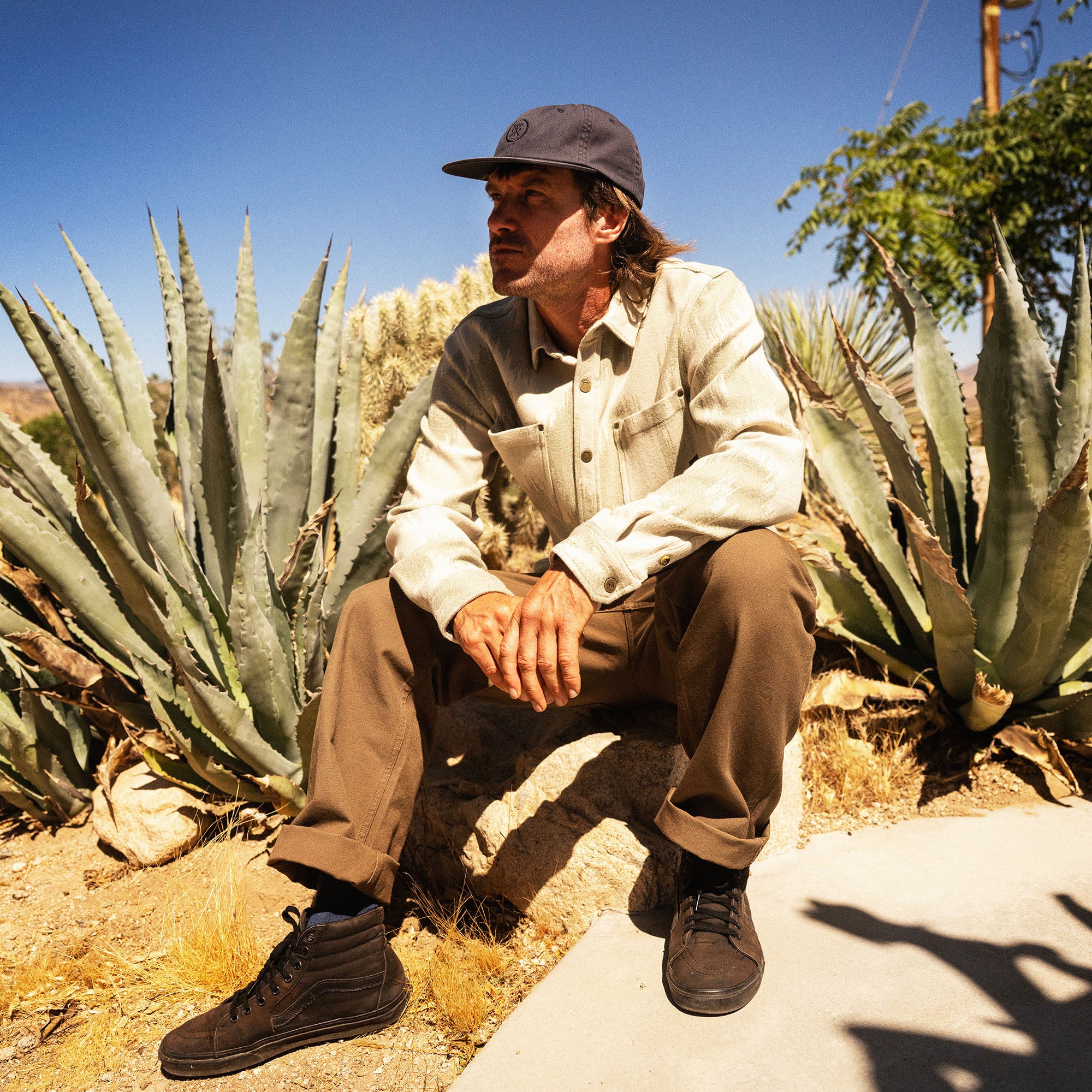 Man wearing cream Journey Jacquard long sleeve flannel shirt and gray baseball cap crouching among tall agave plants in desert landscape