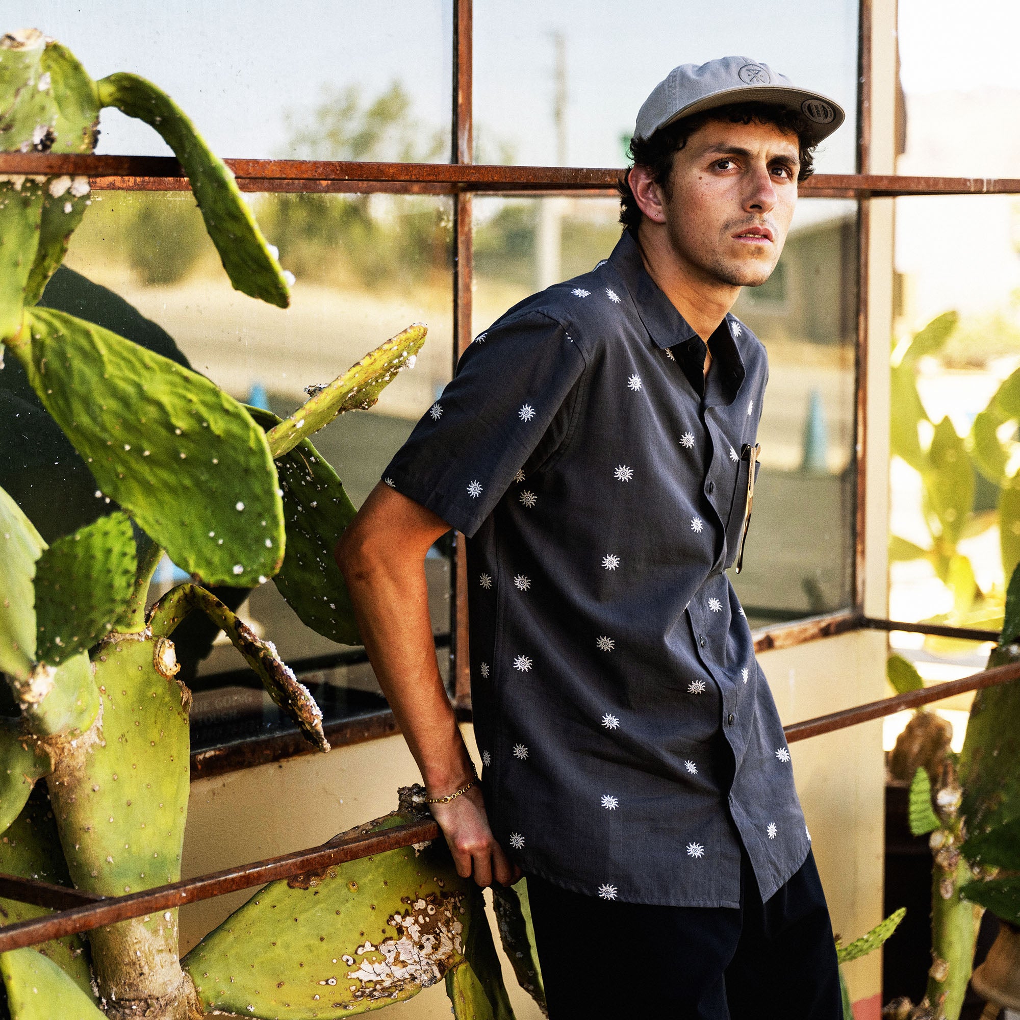 Man wearing charcoal Journey short sleeve button up shirt with white star print, standing in greenhouse with cacti plants