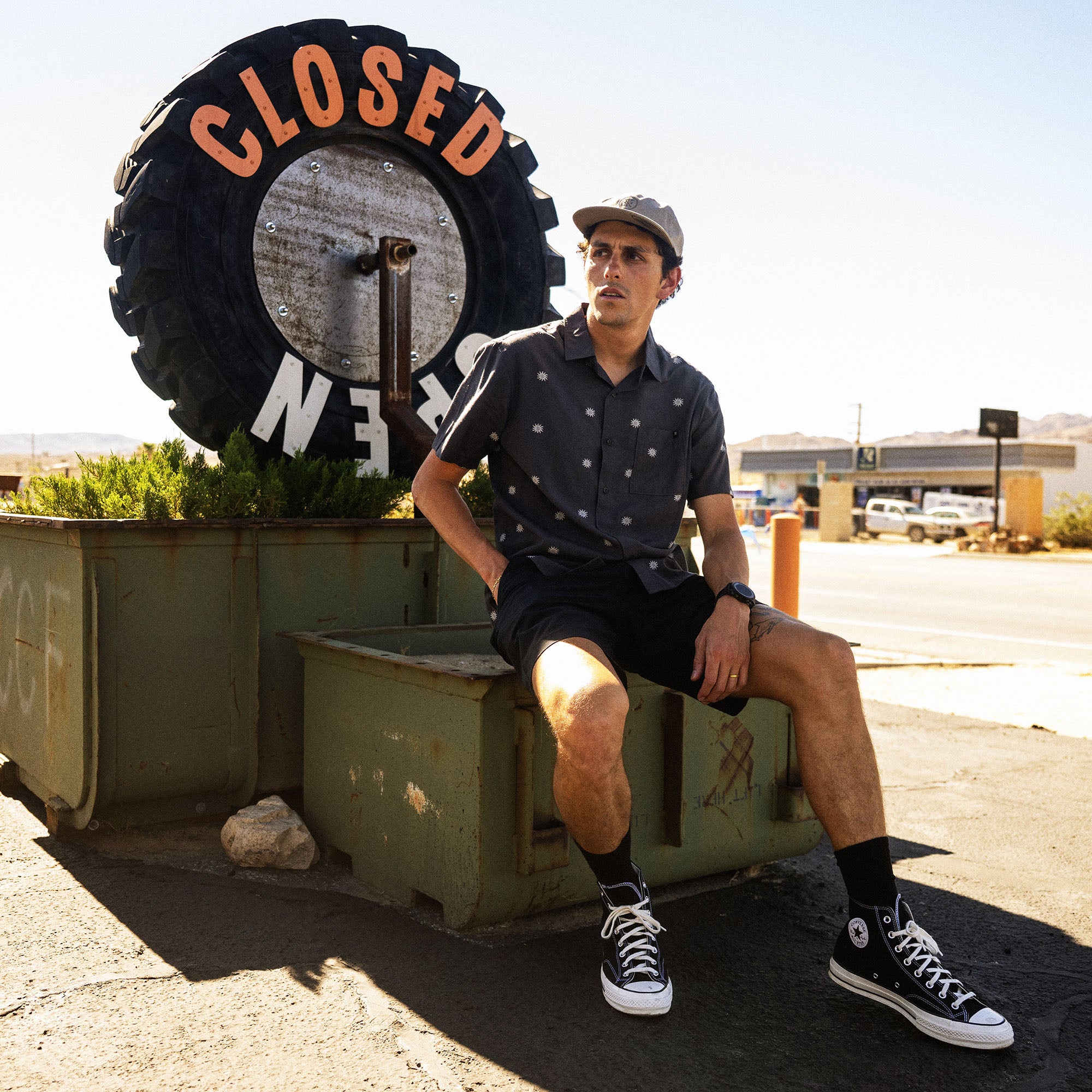 Man in navy patterned short-sleeve shirt and black canvas high-top sneakers sitting outdoors by closed sign
