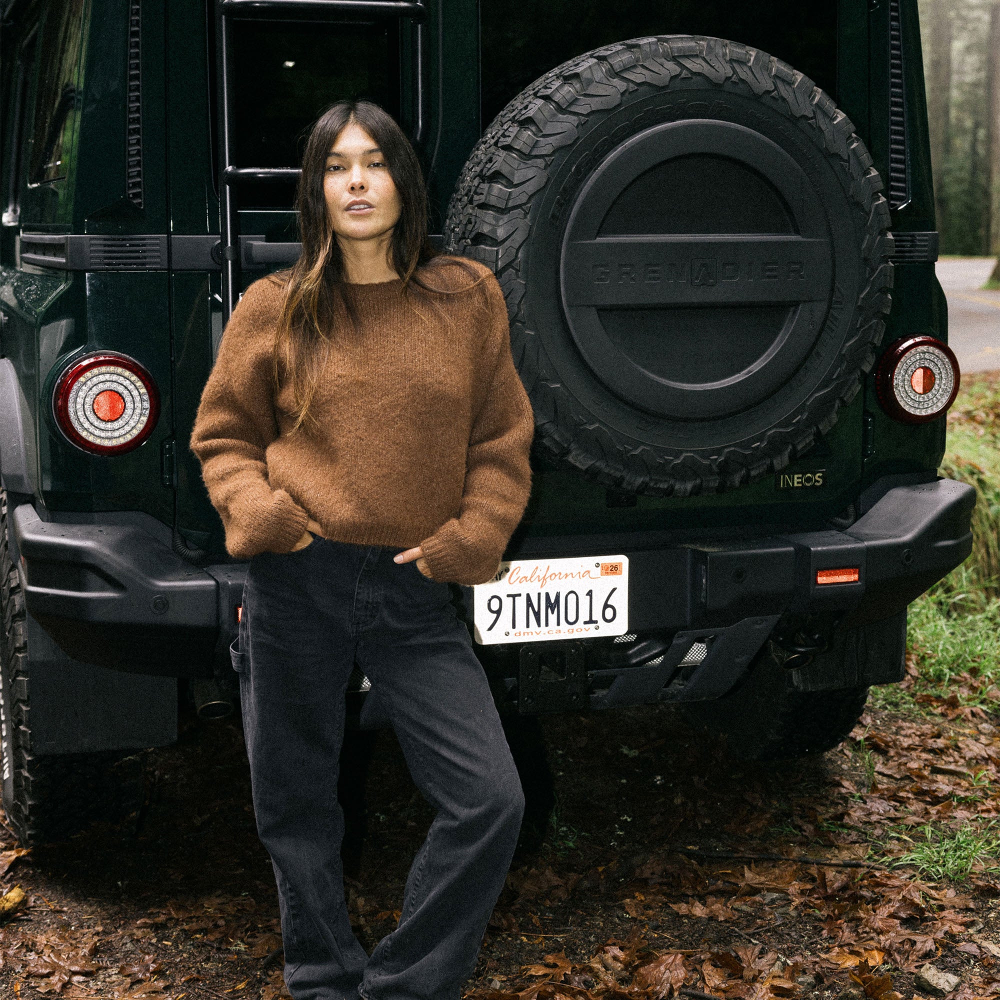 Model wearing a cozy Kos Sweater by Roark styled for a winter adventure, standing beside a snow-covered car fender and bumper, emphasizing warm outdoor fashion.