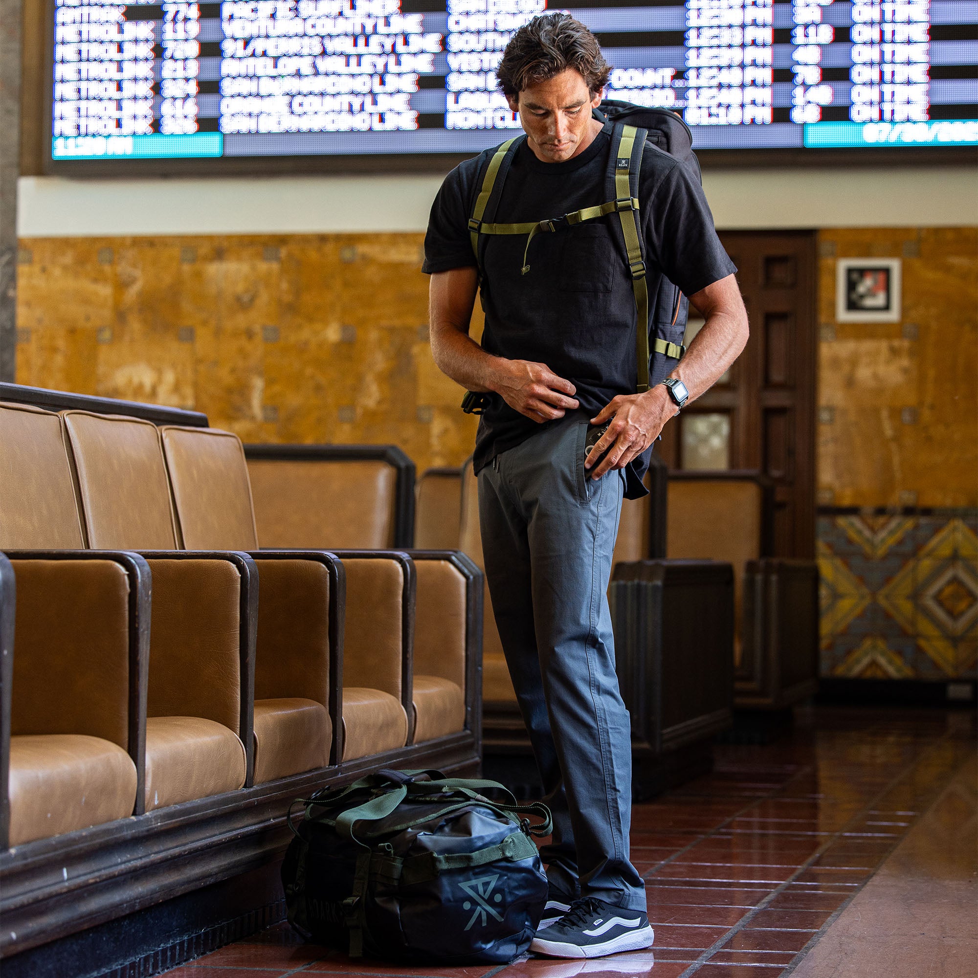 Travel-ready man standing with stylish Layover Pro Pants and an adventure bag, perfectly suited for an active lifestyle.
