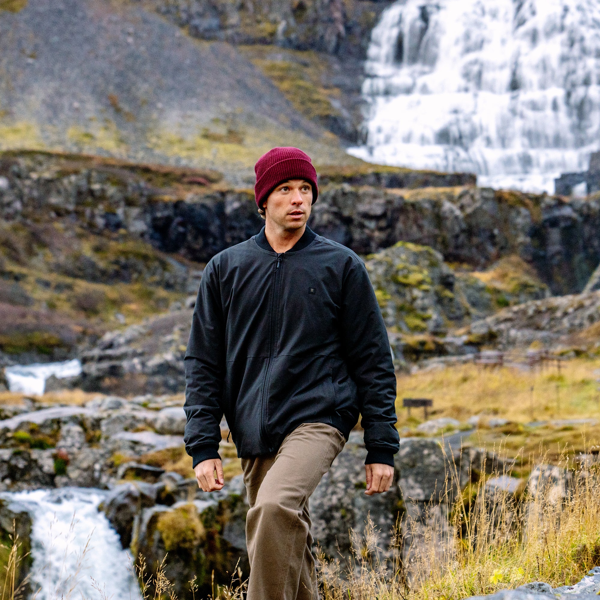 Man wearing black Layover Bomber Jacket and burgundy beanie hiking through mossy rocky terrain with waterfall in background