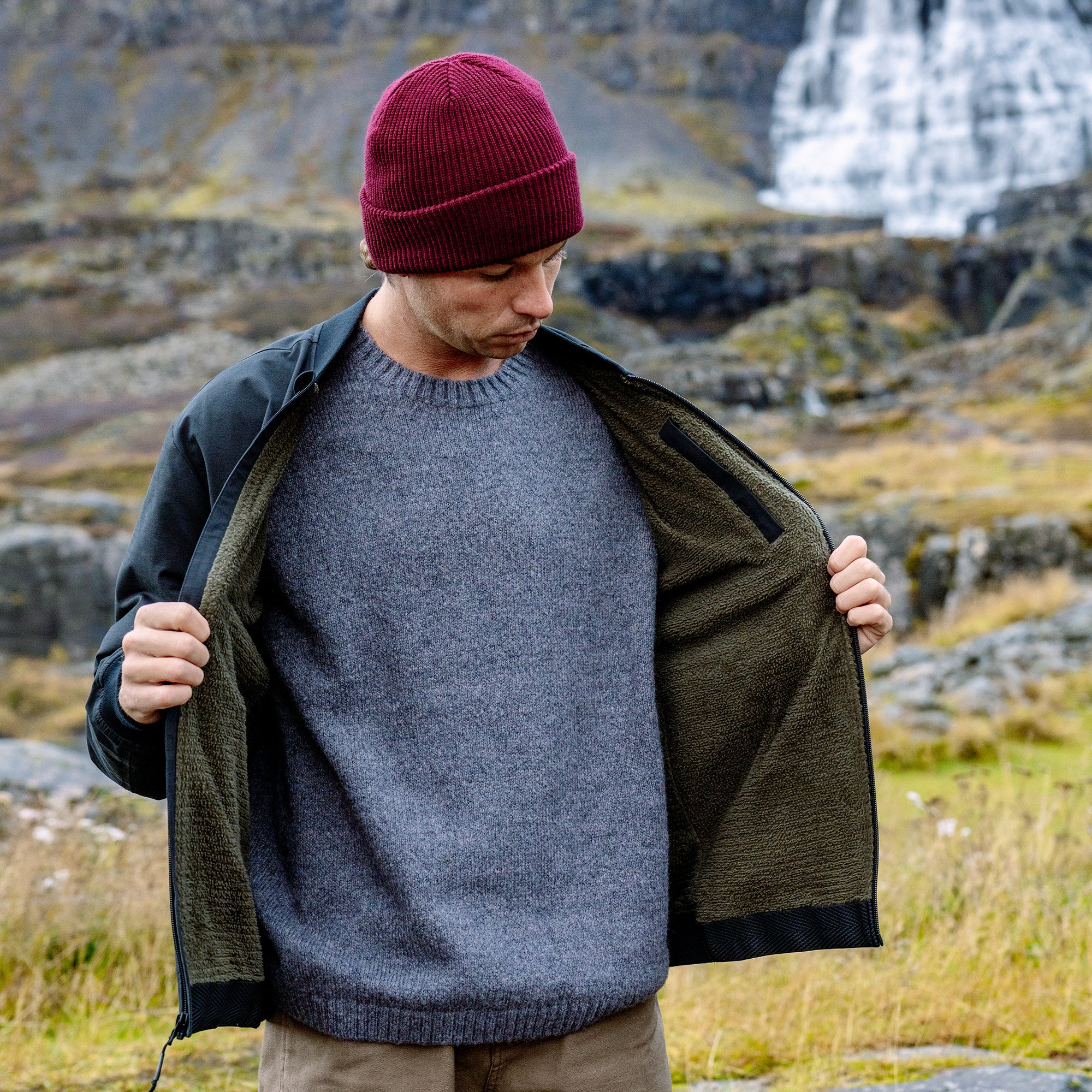 Man wearing burgundy beanie and gray sweater with olive and black layered bomber jacket in rocky mountain landscape