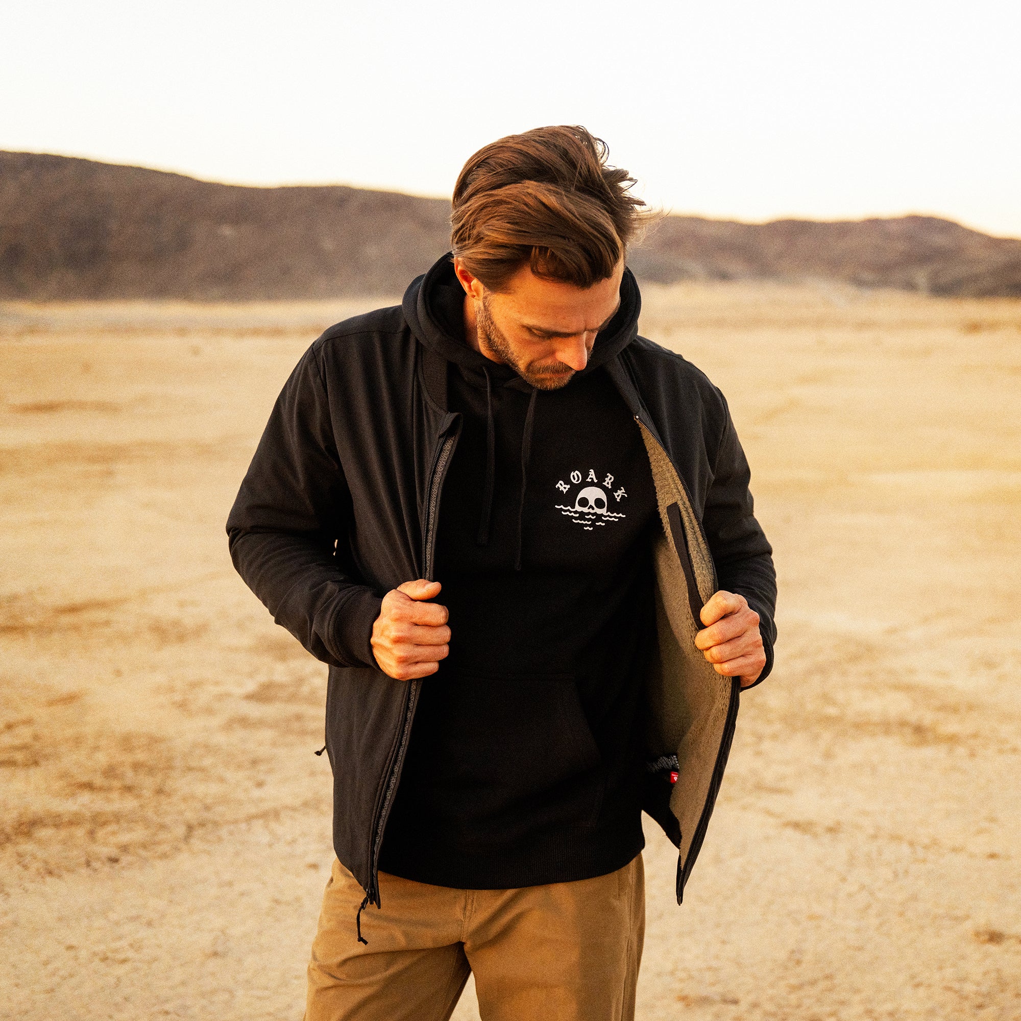 Black layover bomber jacket with skull graphic worn over black hoodie, paired with tan pants in desert setting