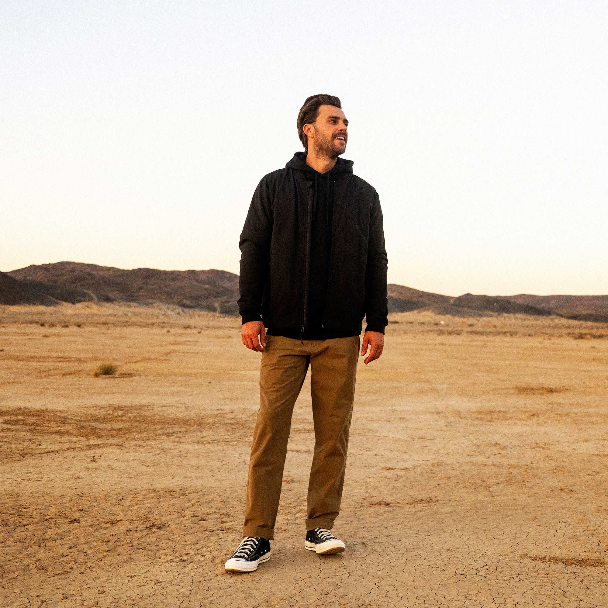 Man wearing black zip-up hoodie and tan chinos with black sneakers standing in desert landscape