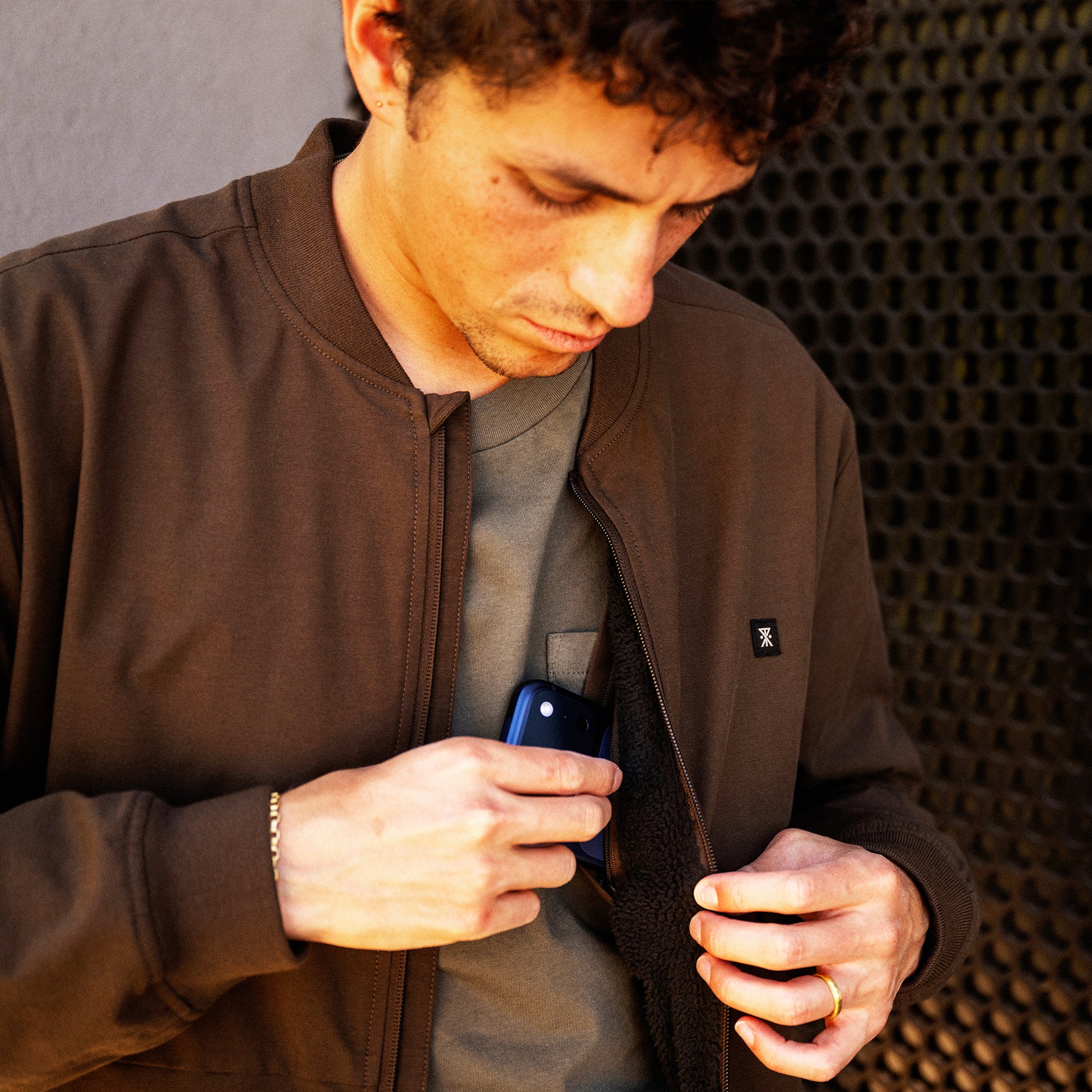 Man wearing brown zip-up bomber jacket over gray shirt, holding smartphone while looking down at screen