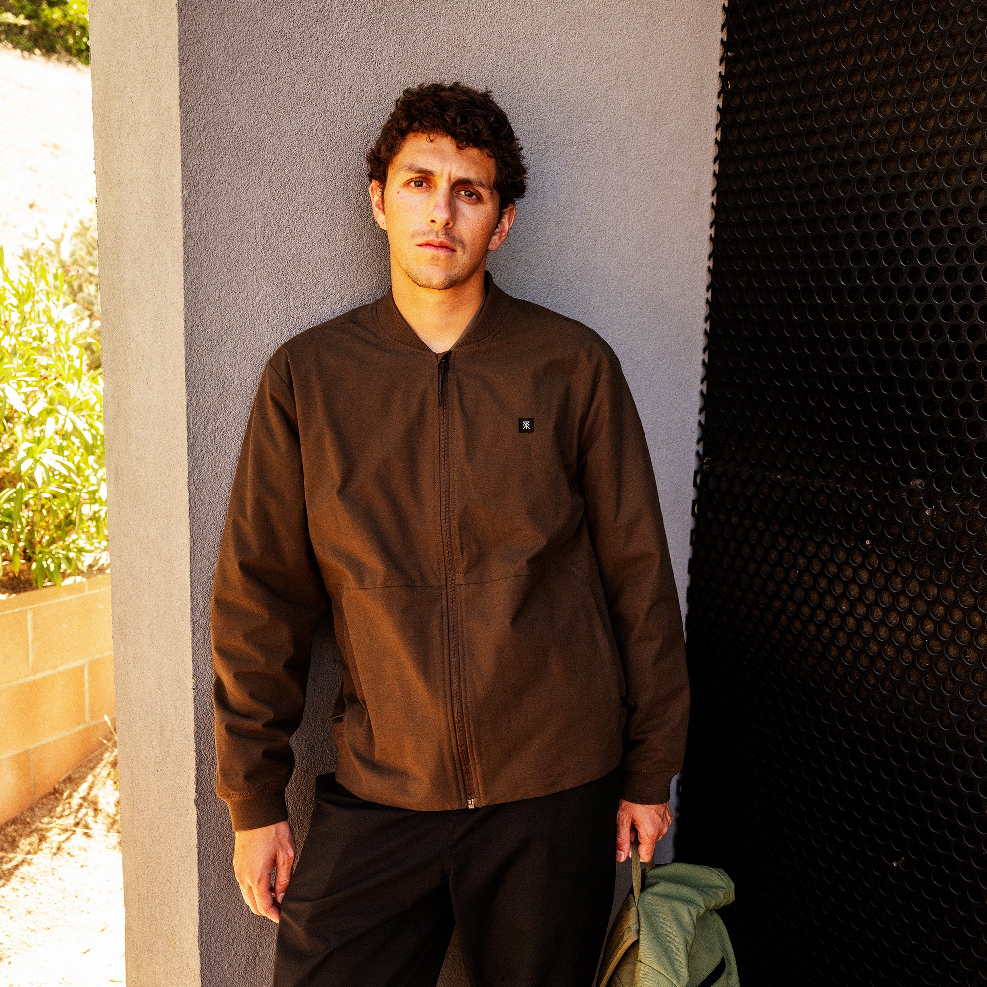 Man wearing brown zip-up bomber jacket with small logo over black pants against grey wall