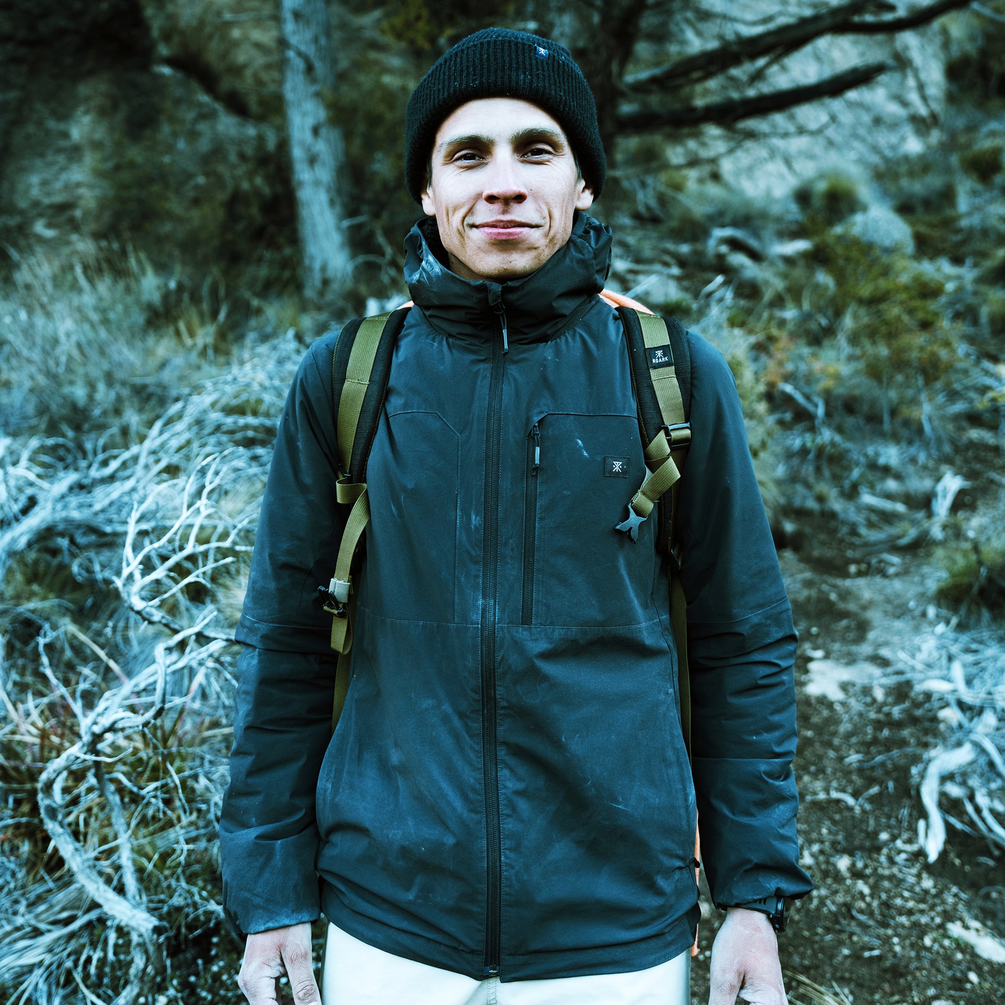 Man wearing black Layover Jacket with black beanie and olive backpack straps in forest setting with frost-covered vegetation