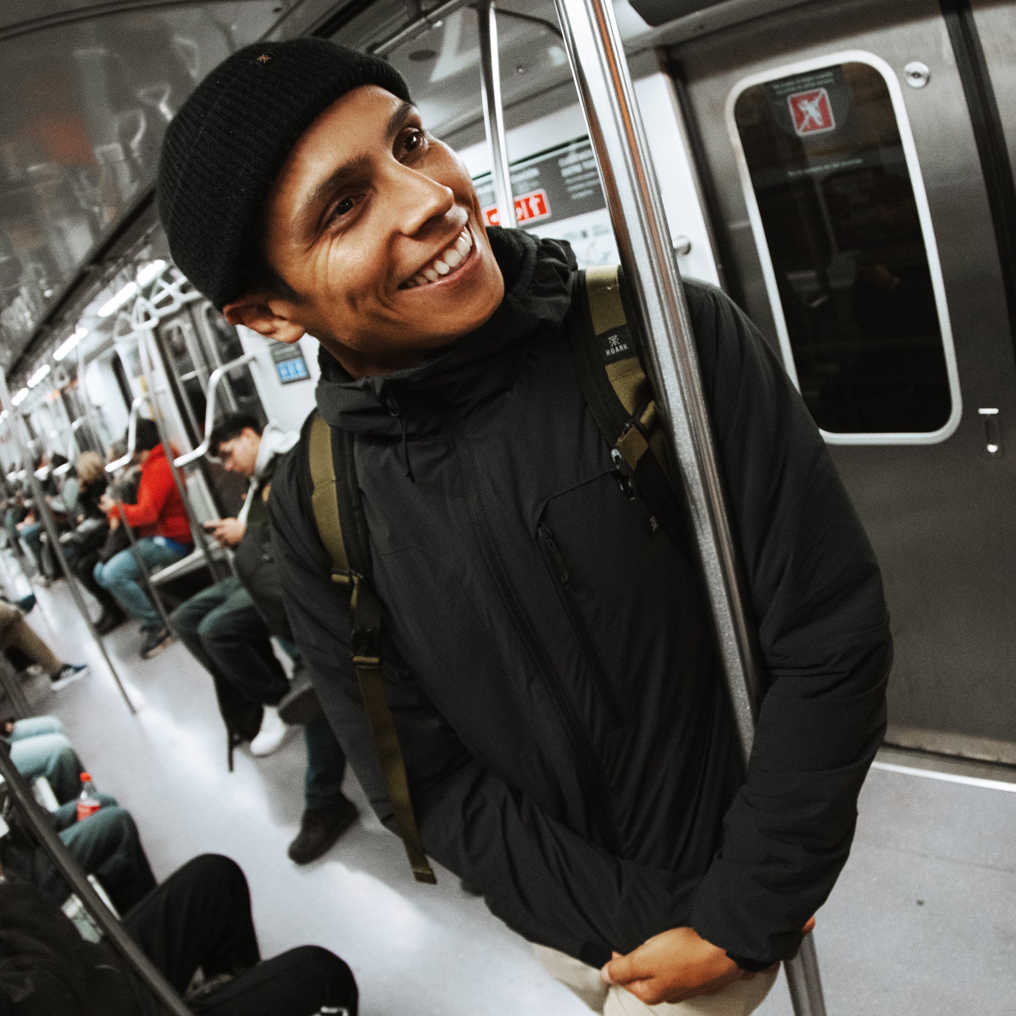 Man wearing black layover jacket with olive shoulder straps smiling inside public transit train car