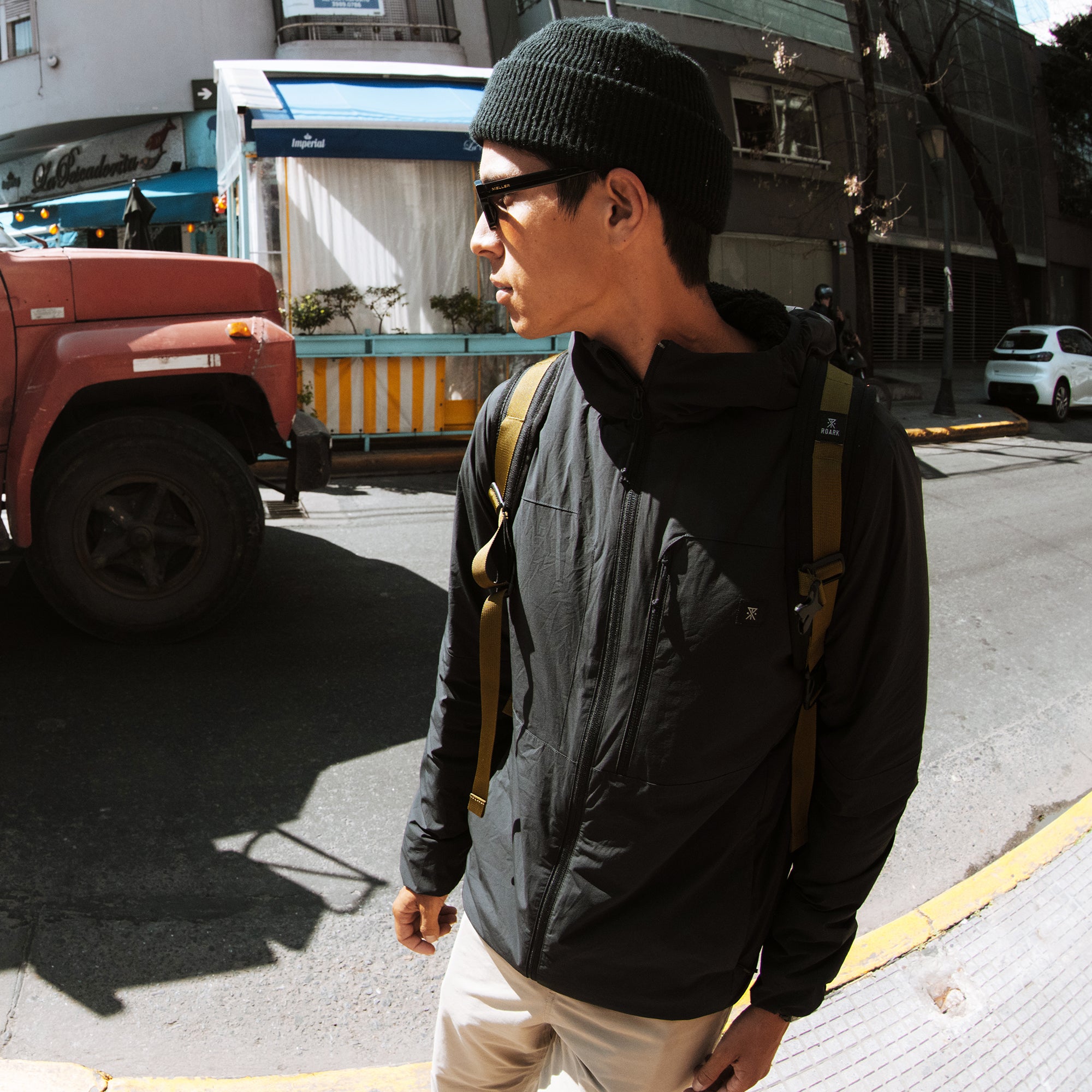 Man wearing black Layover Jacket with tan straps and khaki pants on urban street with parked truck
