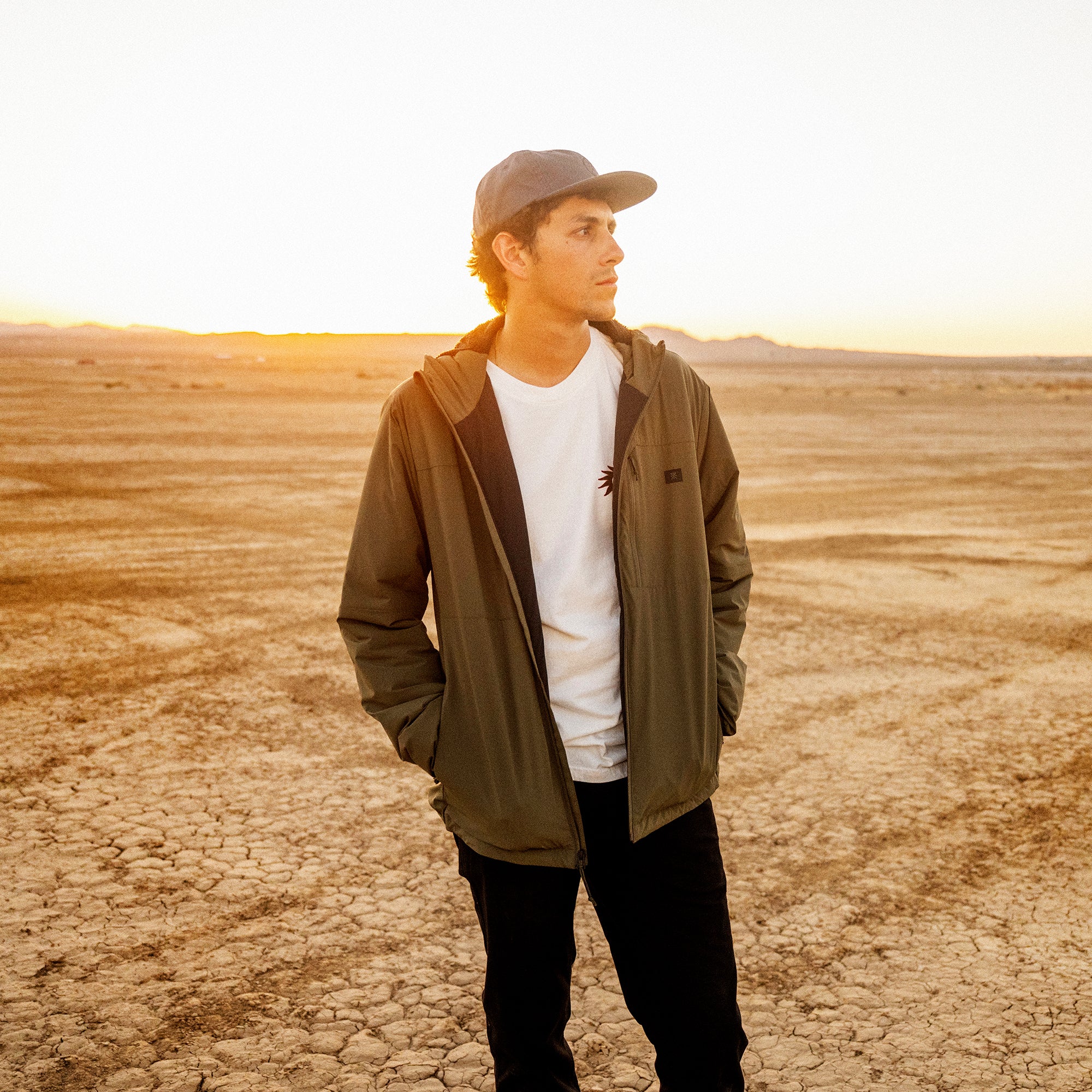 Man wearing olive green Layover Jacket over white t-shirt and black pants in desert landscape at sunset