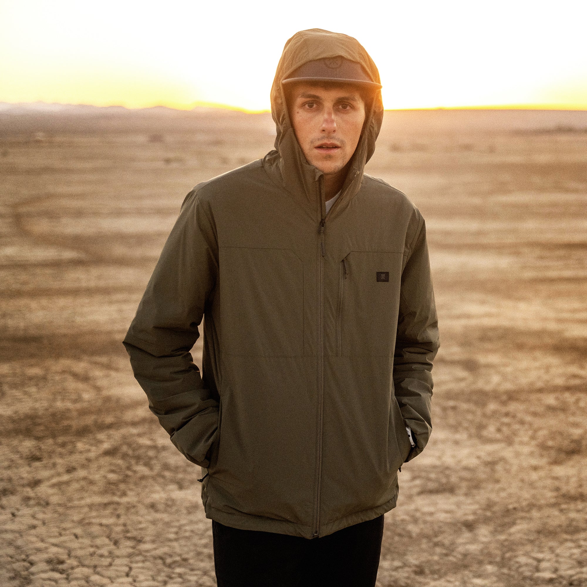 Man wearing olive green hooded zip-up jacket standing in desert landscape at sunset