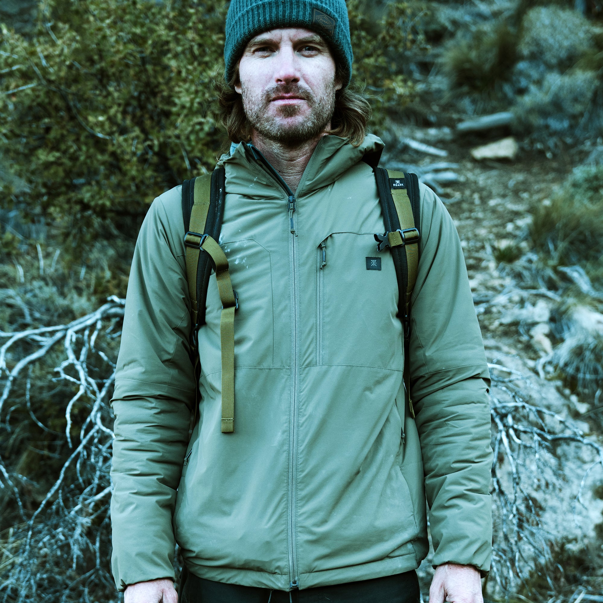 Man wearing sage green Layover Jacket with black backpack straps and teal beanie, standing outdoors in forest setting