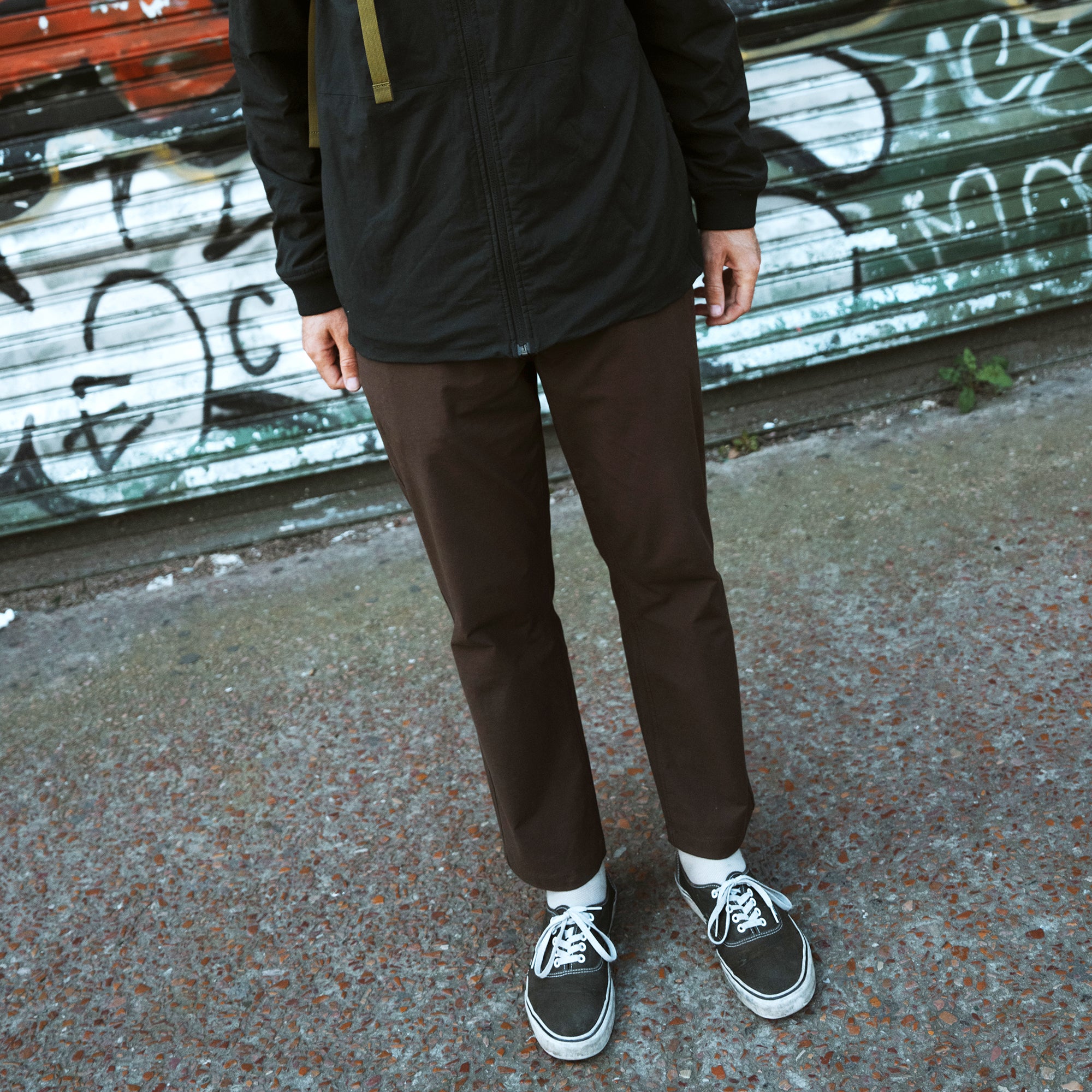 Person wearing brown Layover Pro Traveler Pants with black jacket and white socks with black sneakers standing outdoors