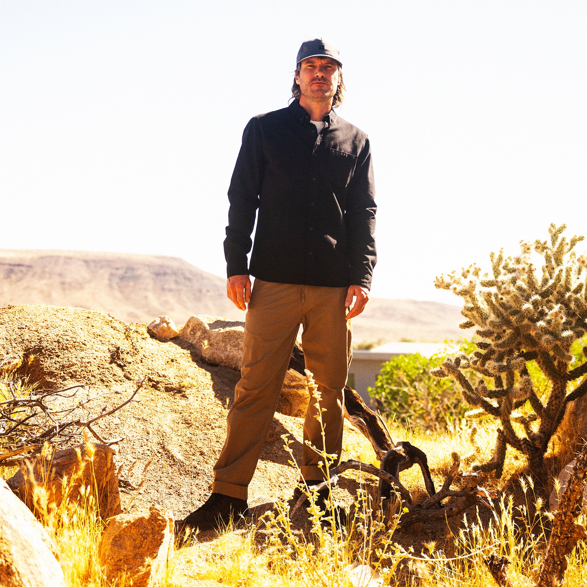 Man wearing black long-sleeve shirt and tan Layover Pro Traveler Pants standing on desert terrain with cacti and mountains