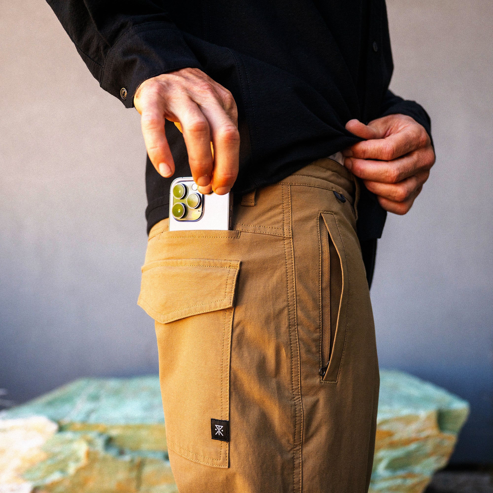 Layover Pro Traveler Pants in dark khaki worn by model holding white smartphone with gold camera, lifestyle shot showing cargo pocket detail