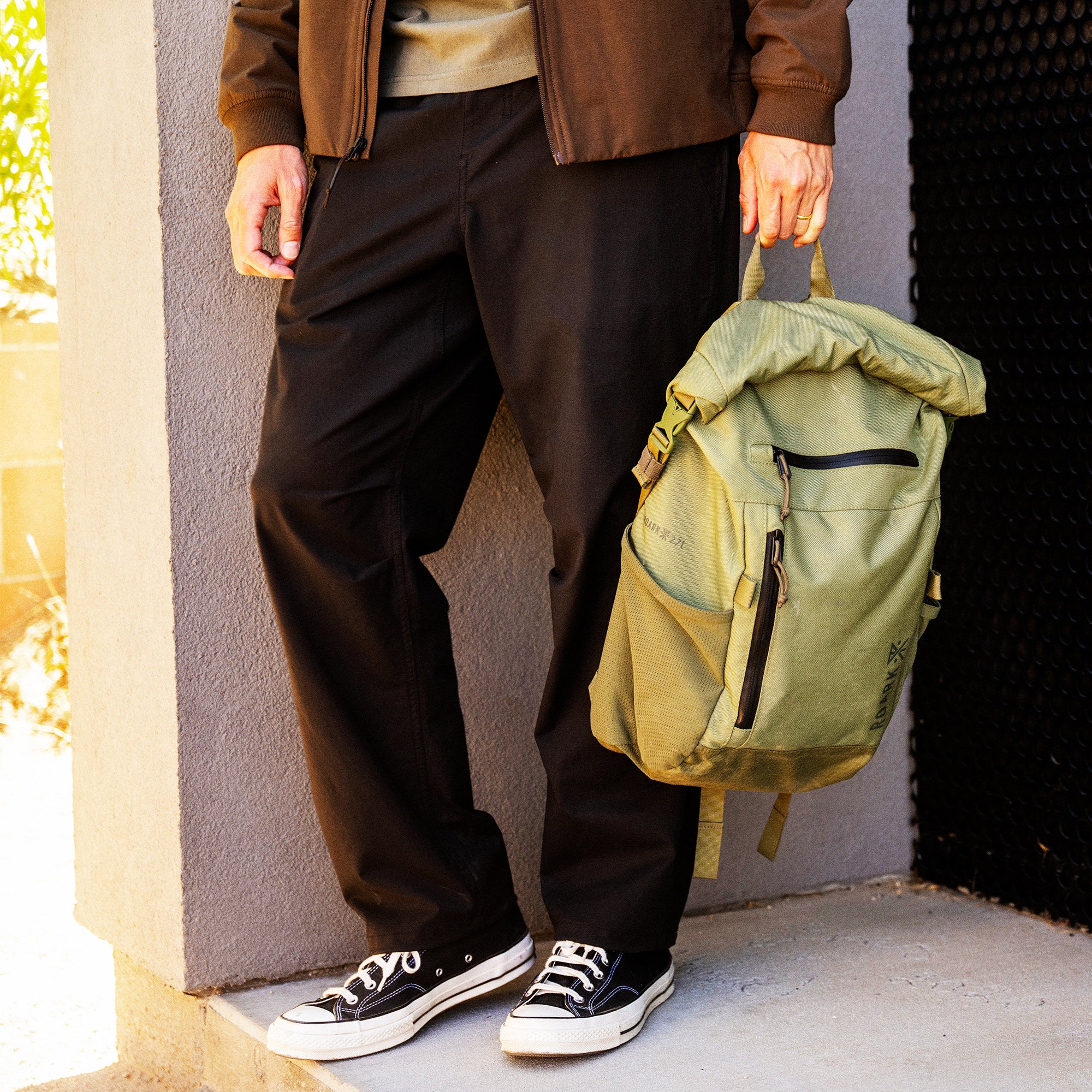 Person wearing brown leather jacket, black pants, and black canvas sneakers holding sage green roll-top backpack with yellow accents