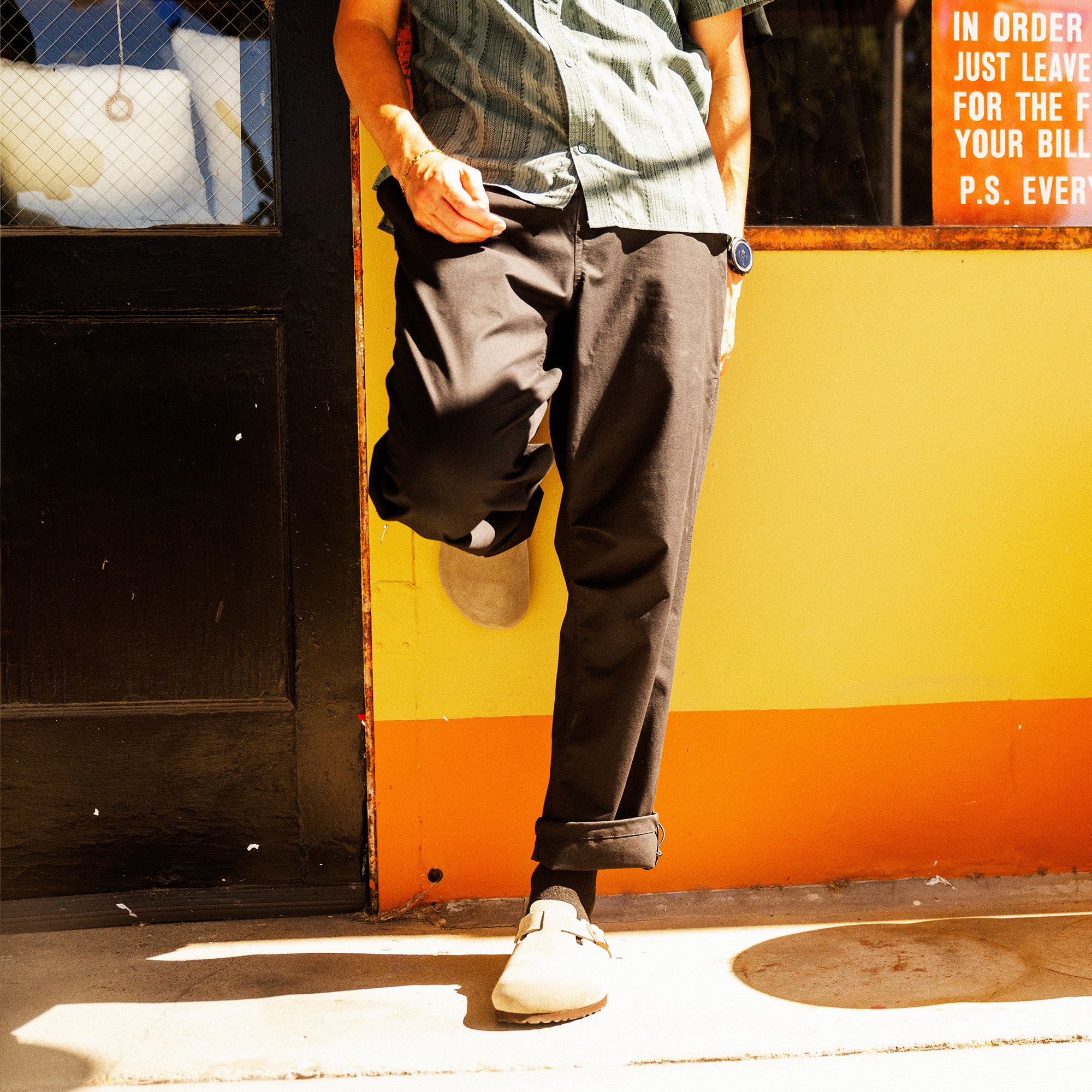 Person wearing charcoal trail pants with rolled cuffs and white canvas sneakers standing against yellow and orange storefront