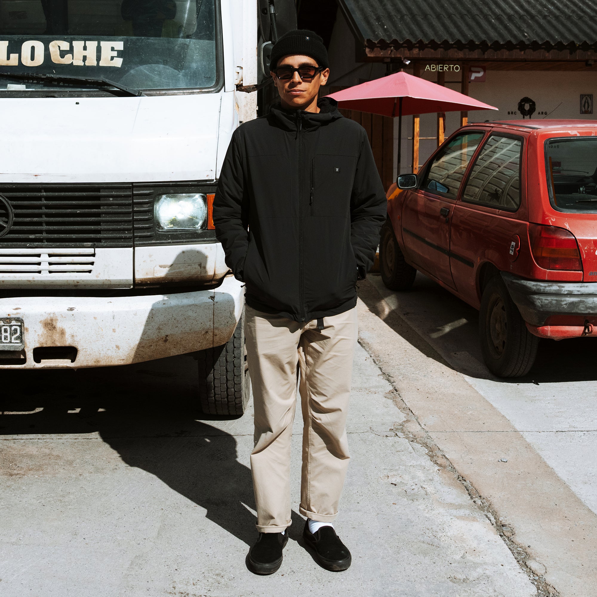 Man wearing black hoodie, beige Layover Trail Pants, black beanie and sunglasses standing in front of parked vehicles