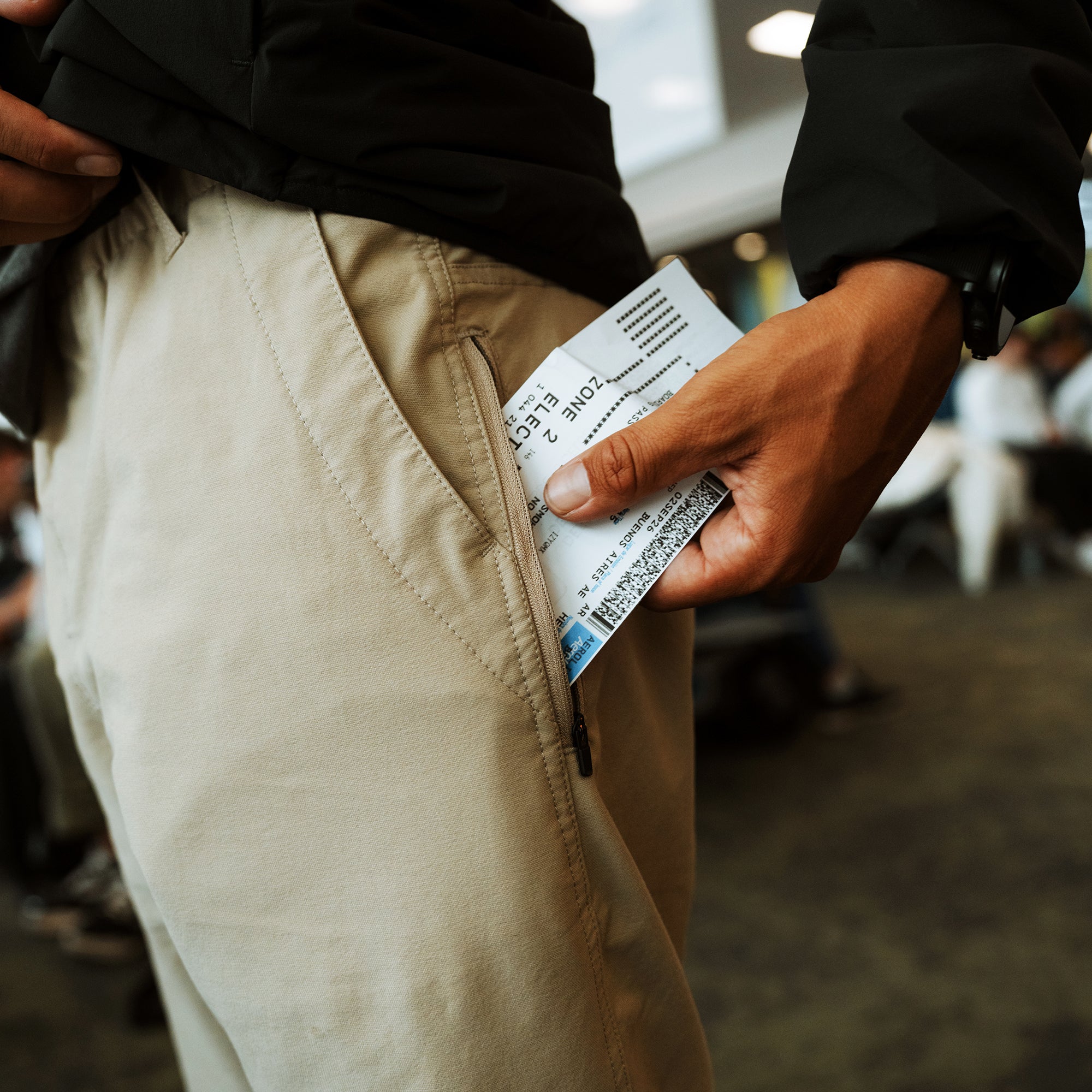 Beige trail pants with side zipper pocket displayed on model wearing black jacket, showing product care label