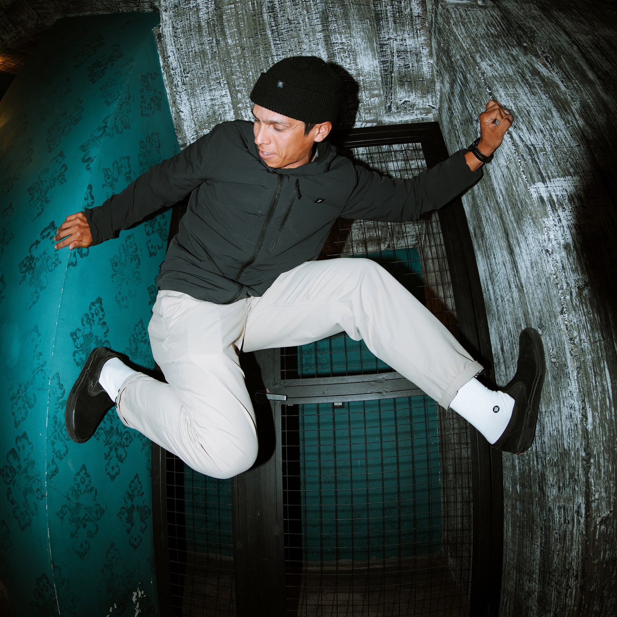 Man wearing black puffer jacket, white Layover Trail Pants, and black beanie jumping in urban setting with turquoise and gray walls