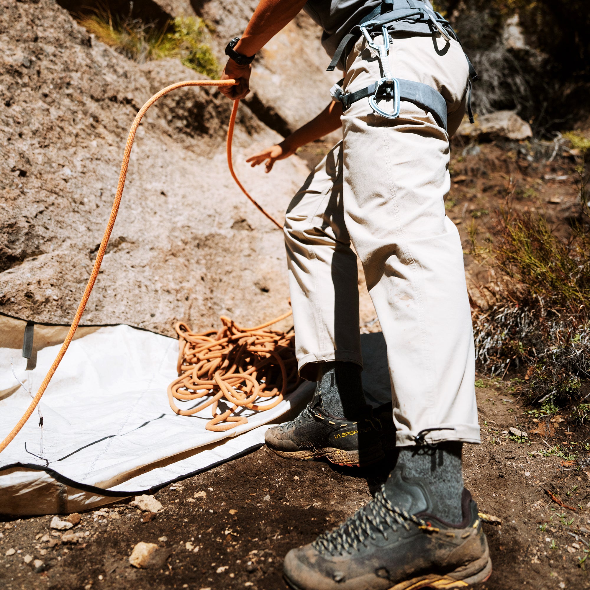 Climber wearing white trail pants with blue harness and hiking boots stands on rocky terrain with orange rope and gear