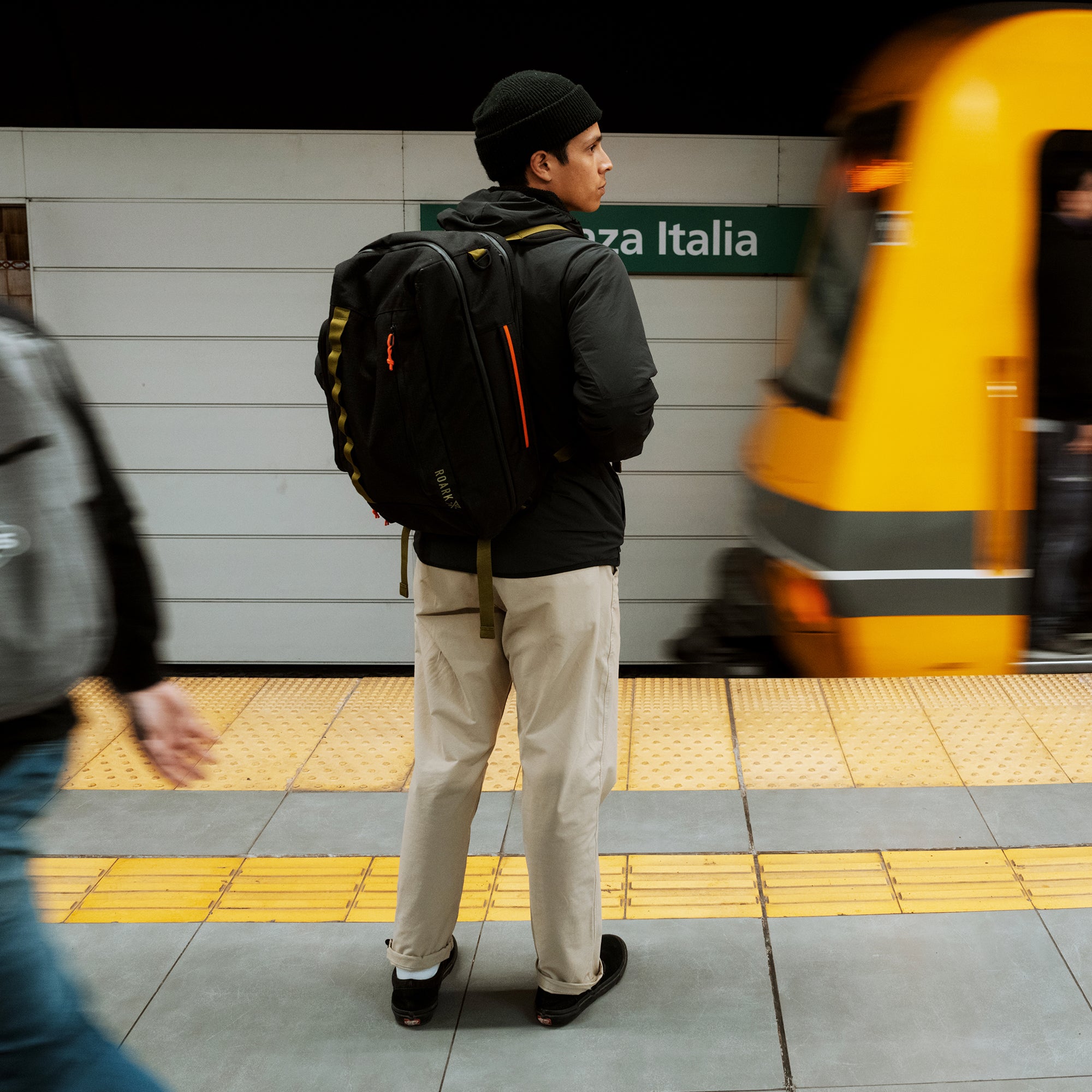 Man wearing black jacket and beige trail pants with black backpack waiting at transit station platform