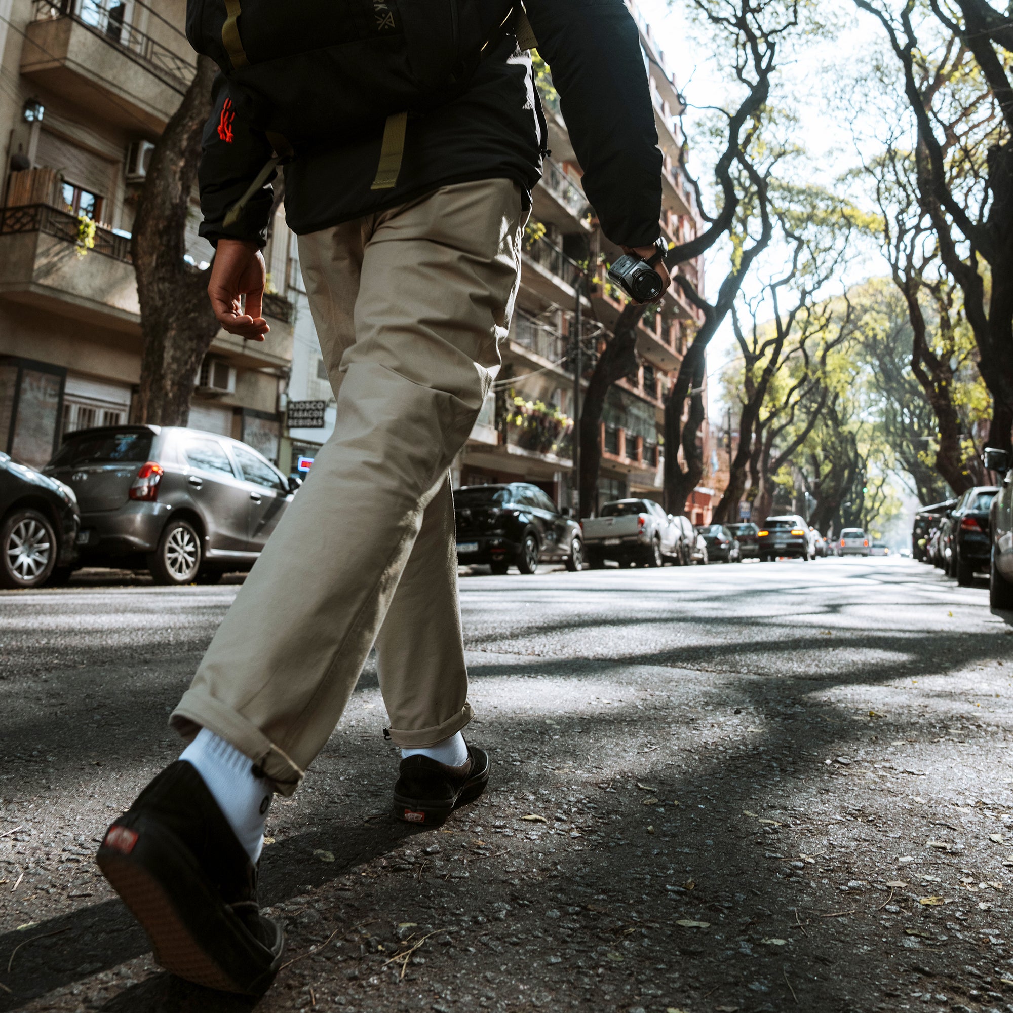 Person wearing khaki Layover Trail Pants with white socks and dark sandals walking down a tree-lined urban street with parked cars and