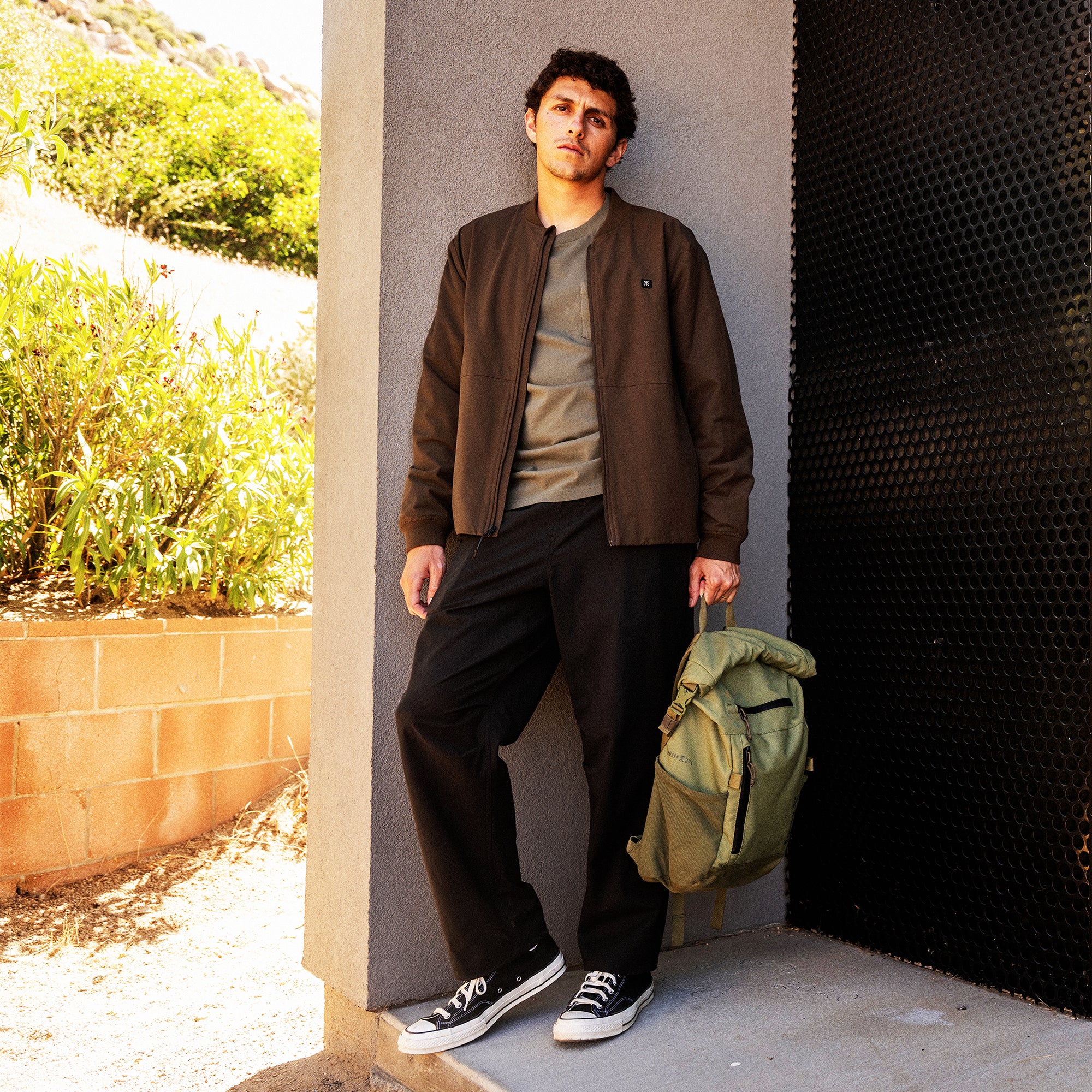 Man wearing brown bomber jacket, gray t-shirt, black Layover Trail Pants, and black sneakers, holding olive backpack against concrete wall