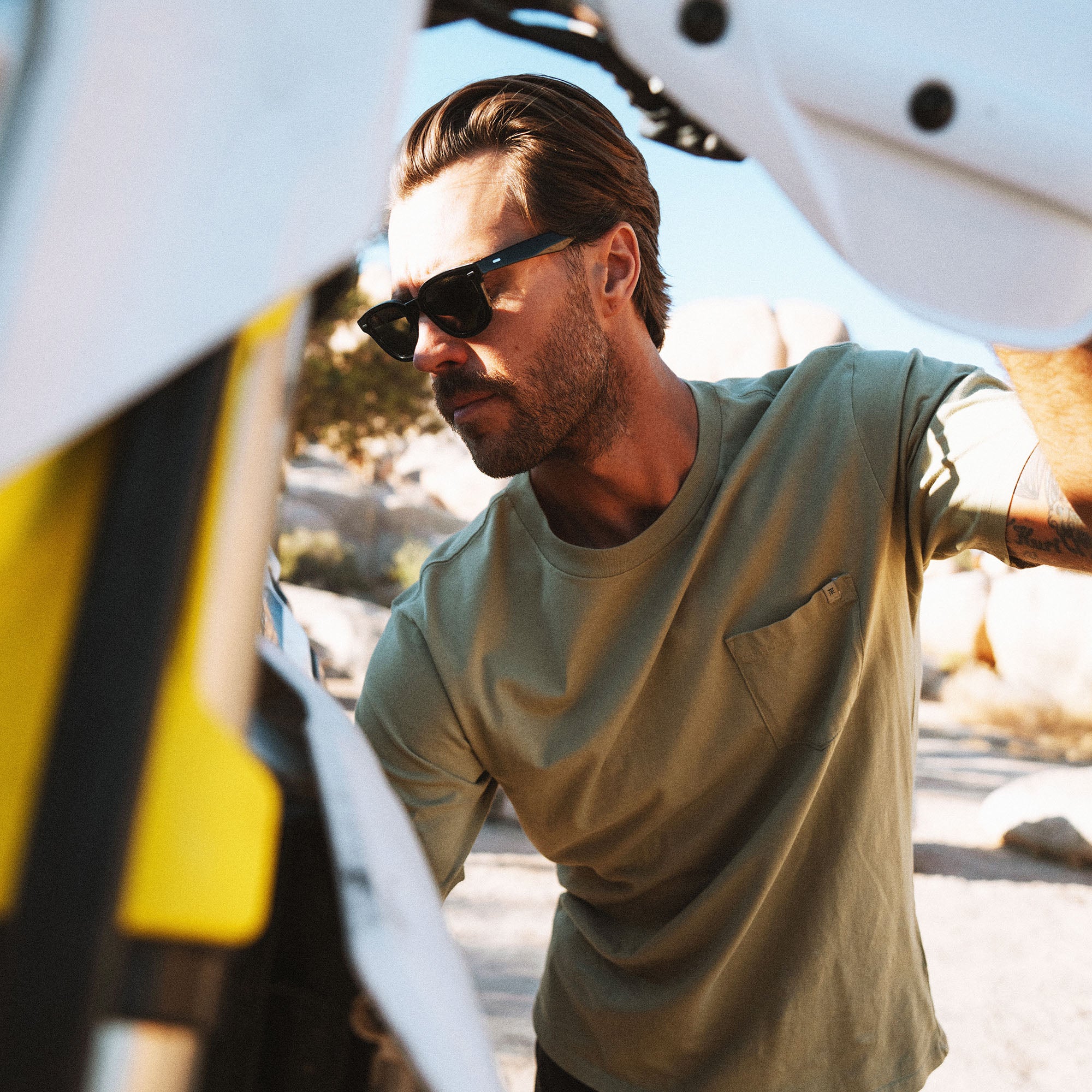 Man wearing sage green crew neck t-shirt and black sunglasses standing outdoors near white boat