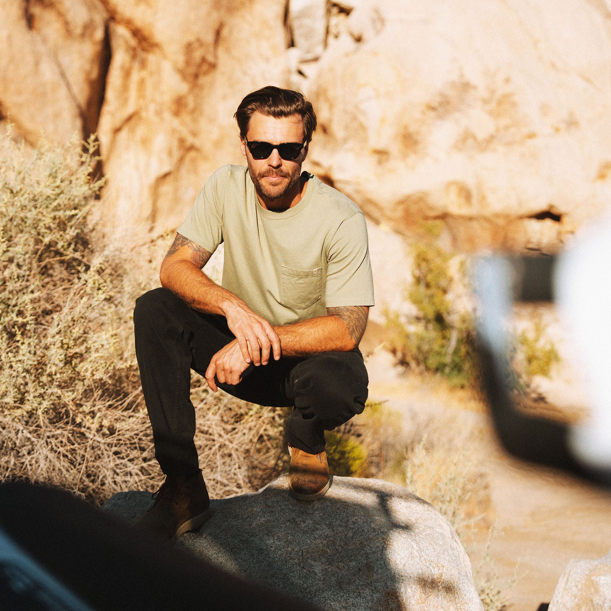 Man wearing washed dusty sage short sleeve tee with chest pocket and black sunglasses, crouching outdoors against stone wall