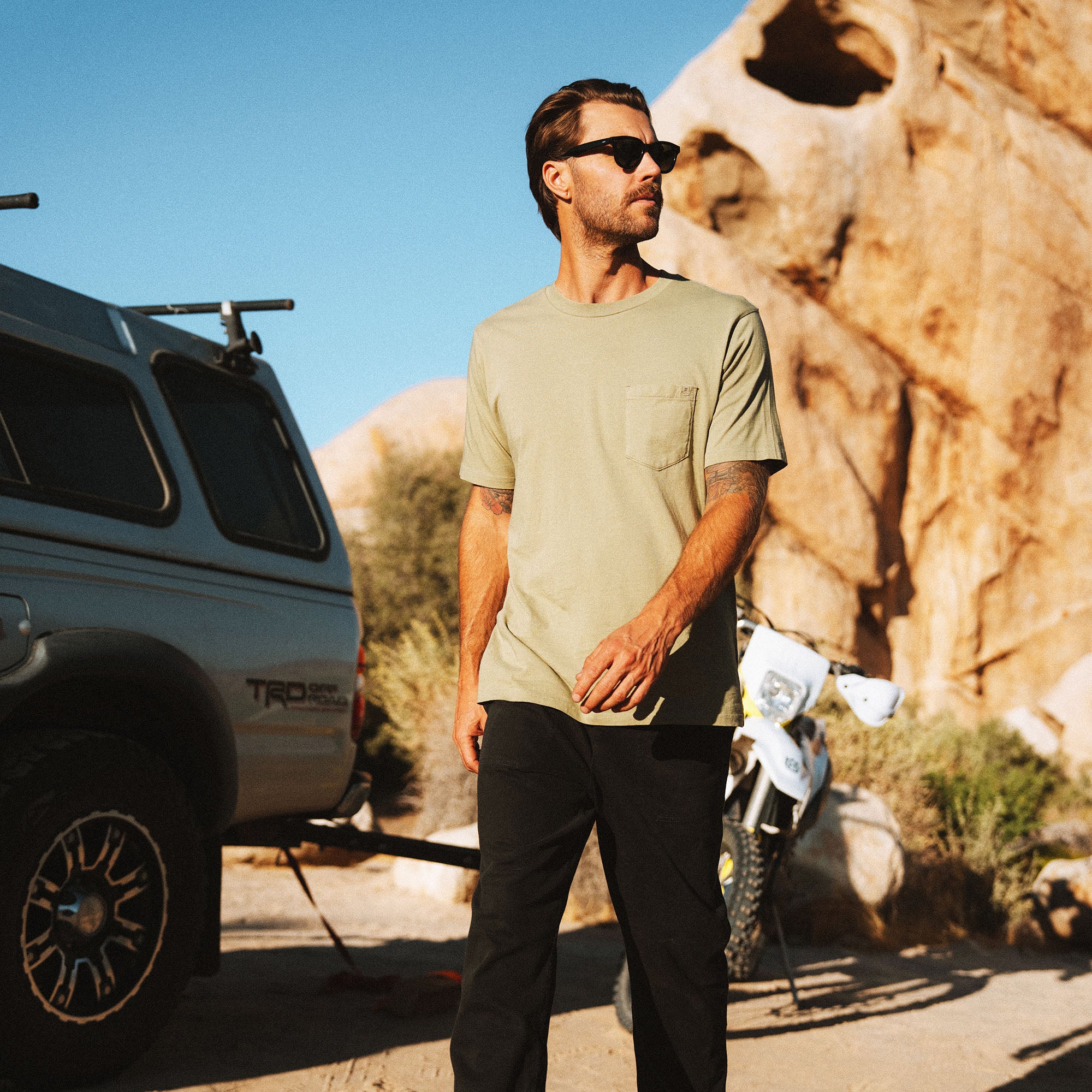 Man wearing sage green pocket t-shirt and black pants with sunglasses standing near desert rocks and vehicle