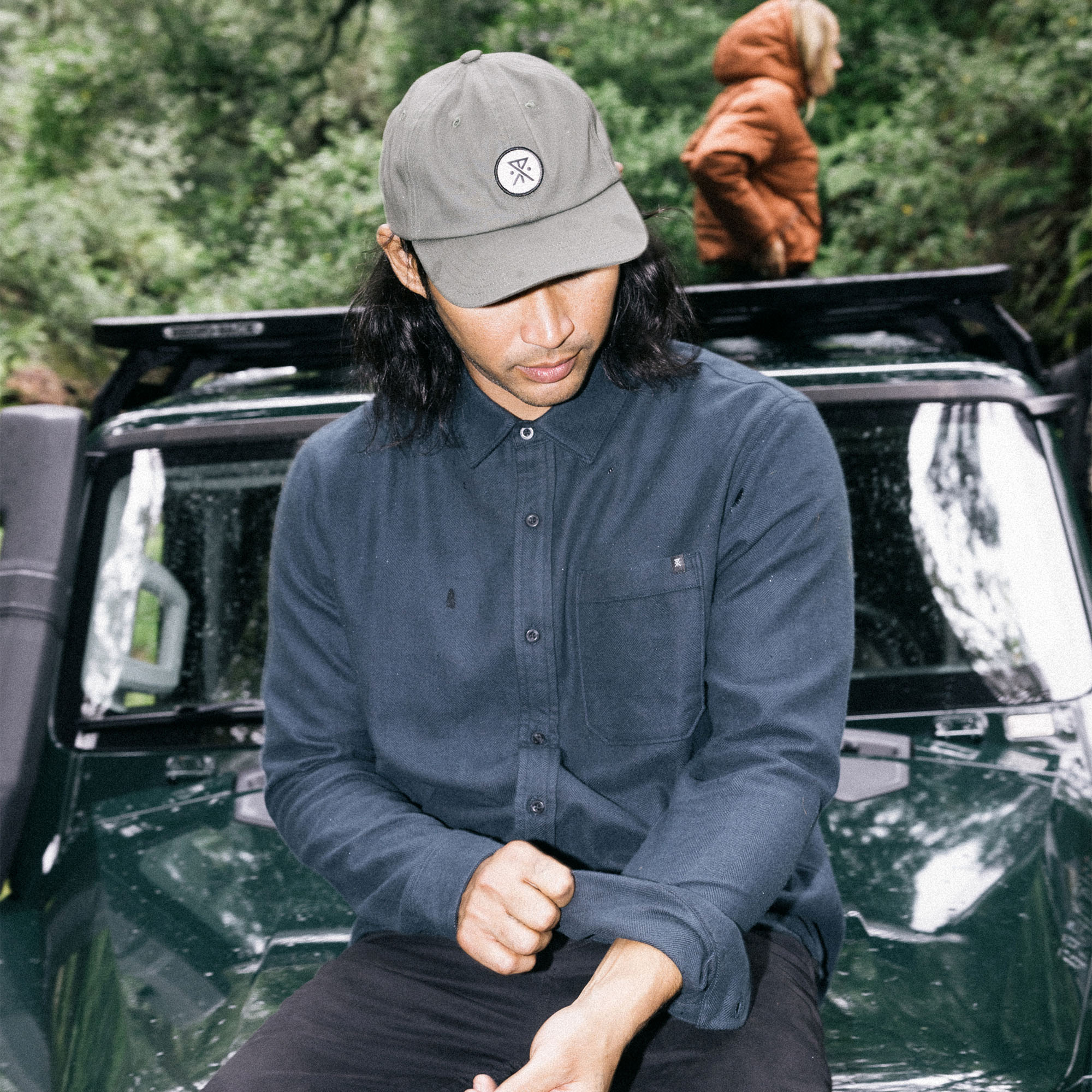 Man in a 'Made to Fade' long sleeve button-up shirt by Roark, stylishly accessorized with a baseball cap, next to a car door, ready for a road trip adventure.