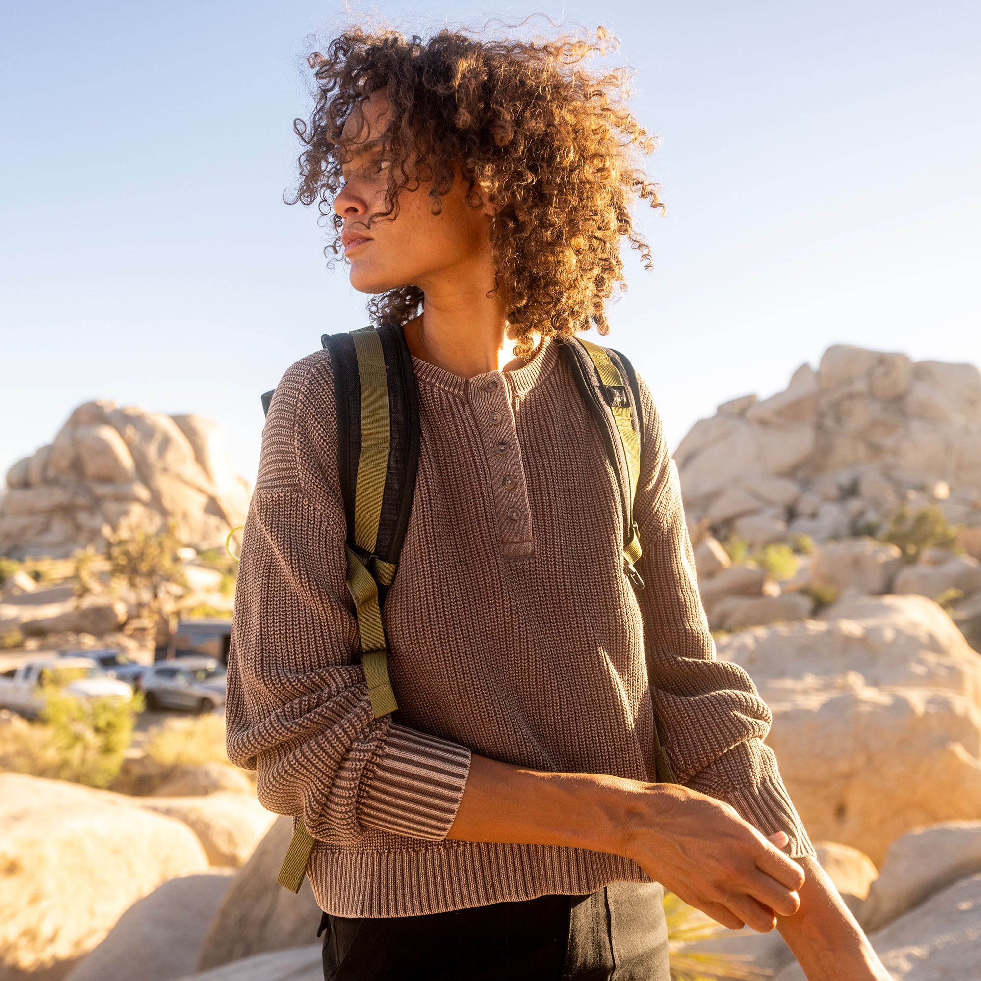 Woman wearing taupe Made To Fade long sleeve sweater with backpack standing in desert rocky landscape at sunset