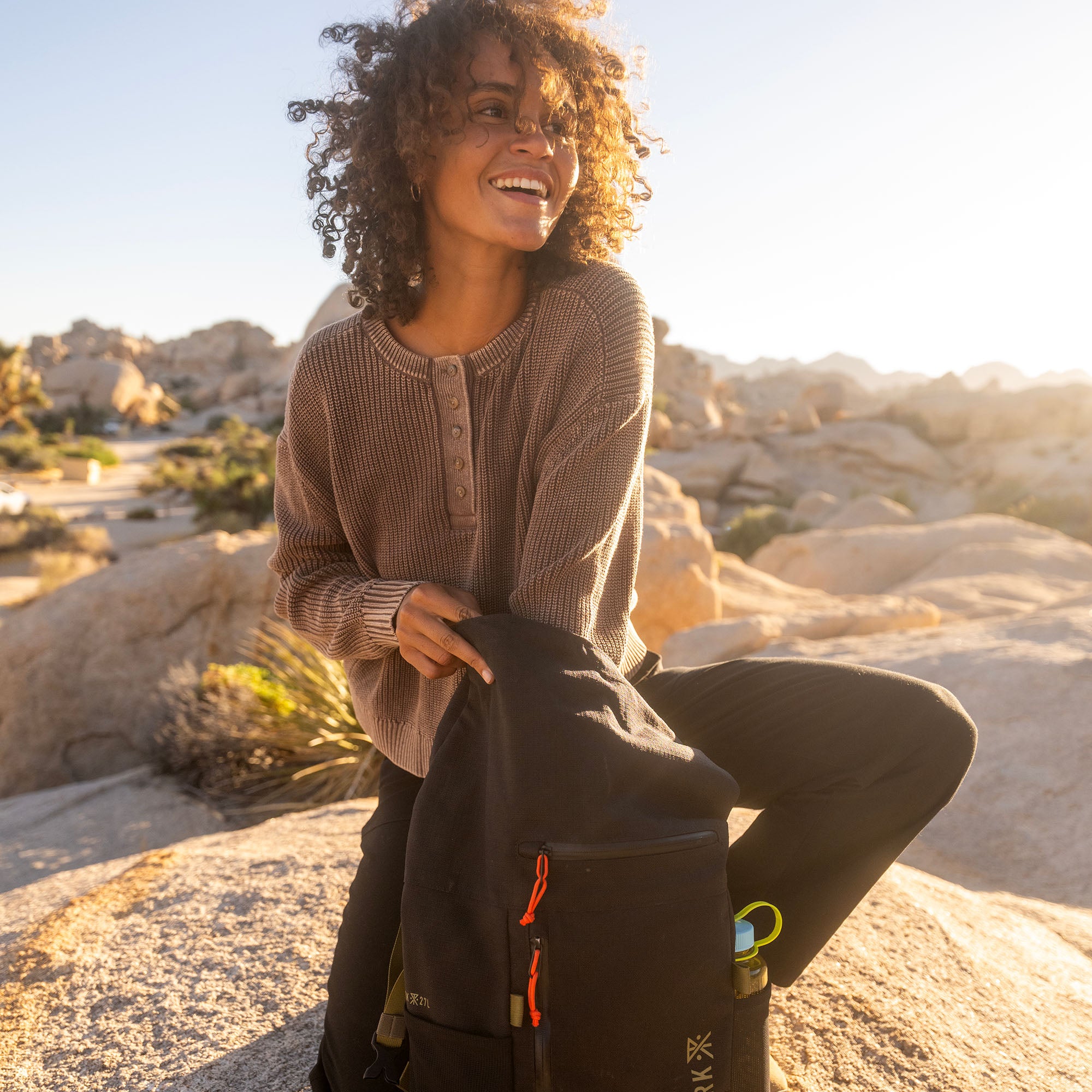 Woman wearing beige ribbed long-sleeve henley sweater sitting outdoors with rocky desert landscape in golden sunlight
