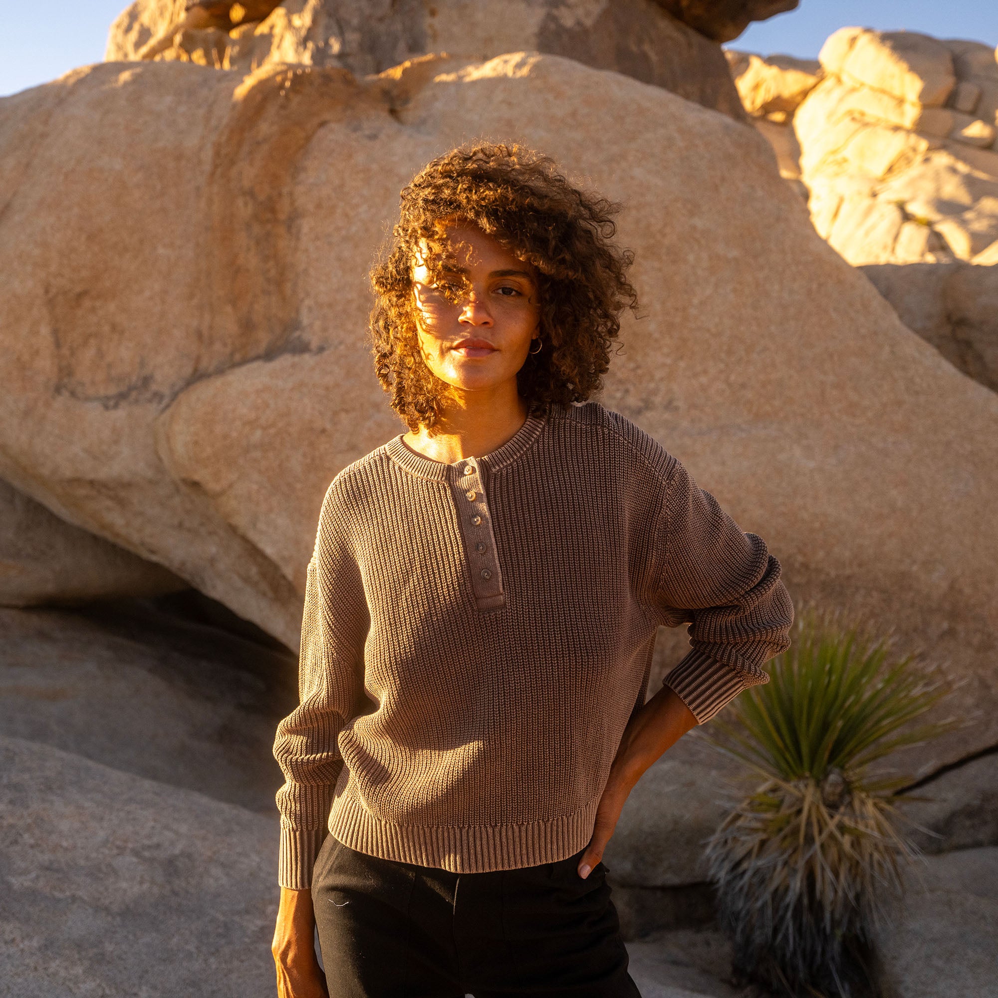 Coco Bean long sleeve sweater with henley neckline on model standing outdoors among large rocks at sunset