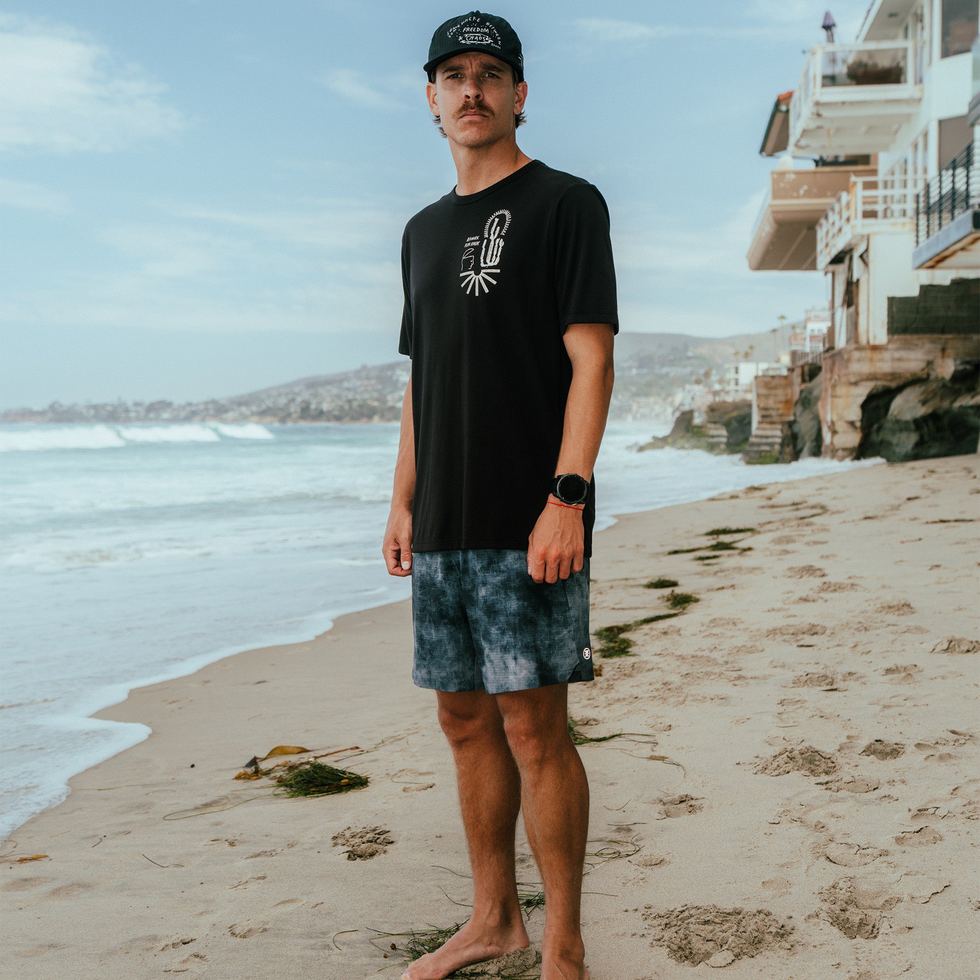 Man wearing black short sleeve tee with graphic design and blue tie-dye shorts standing on beach
