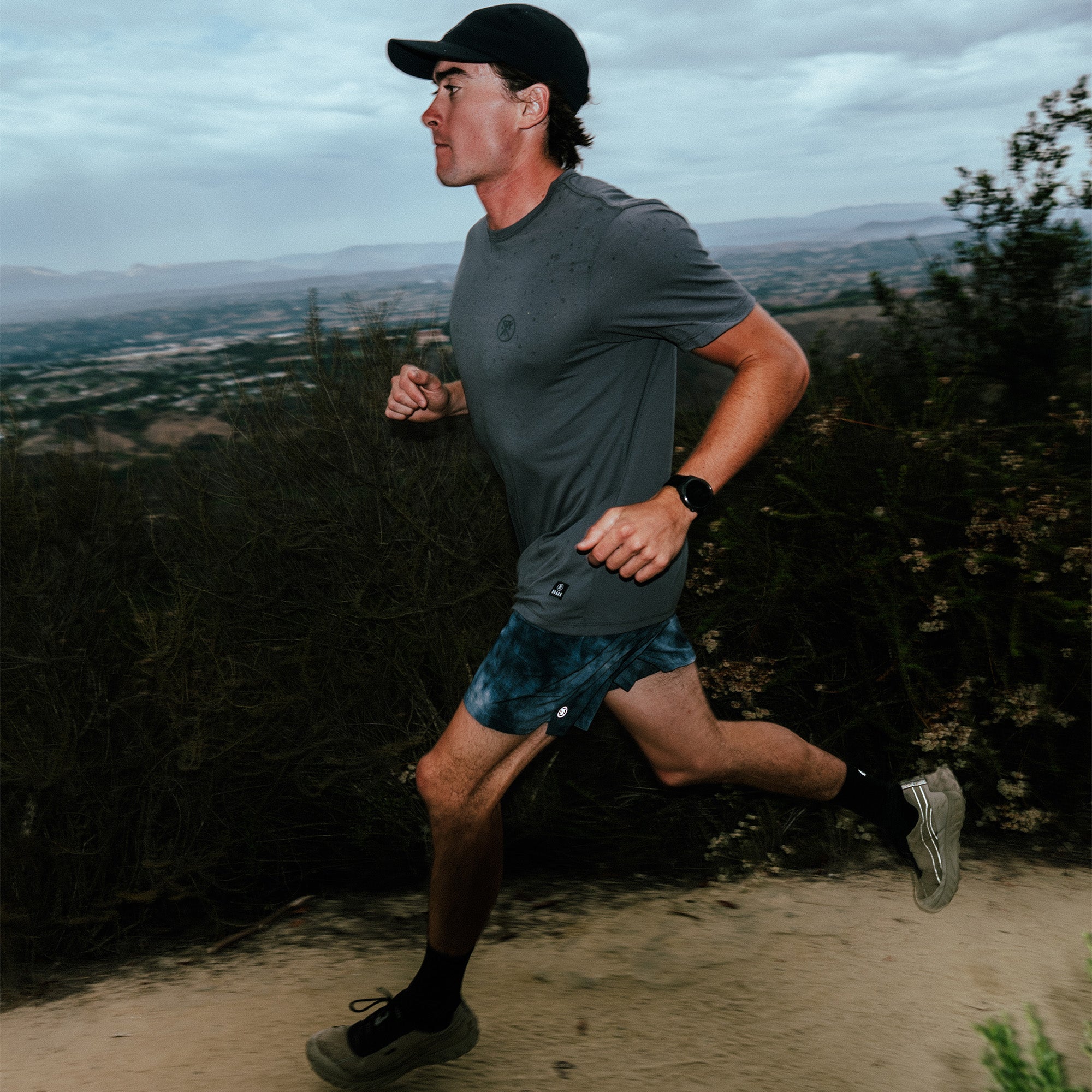 Man wearing charcoal Mathis Active Short Sleeve Tee while trail running on hillside with valley landscape behind him