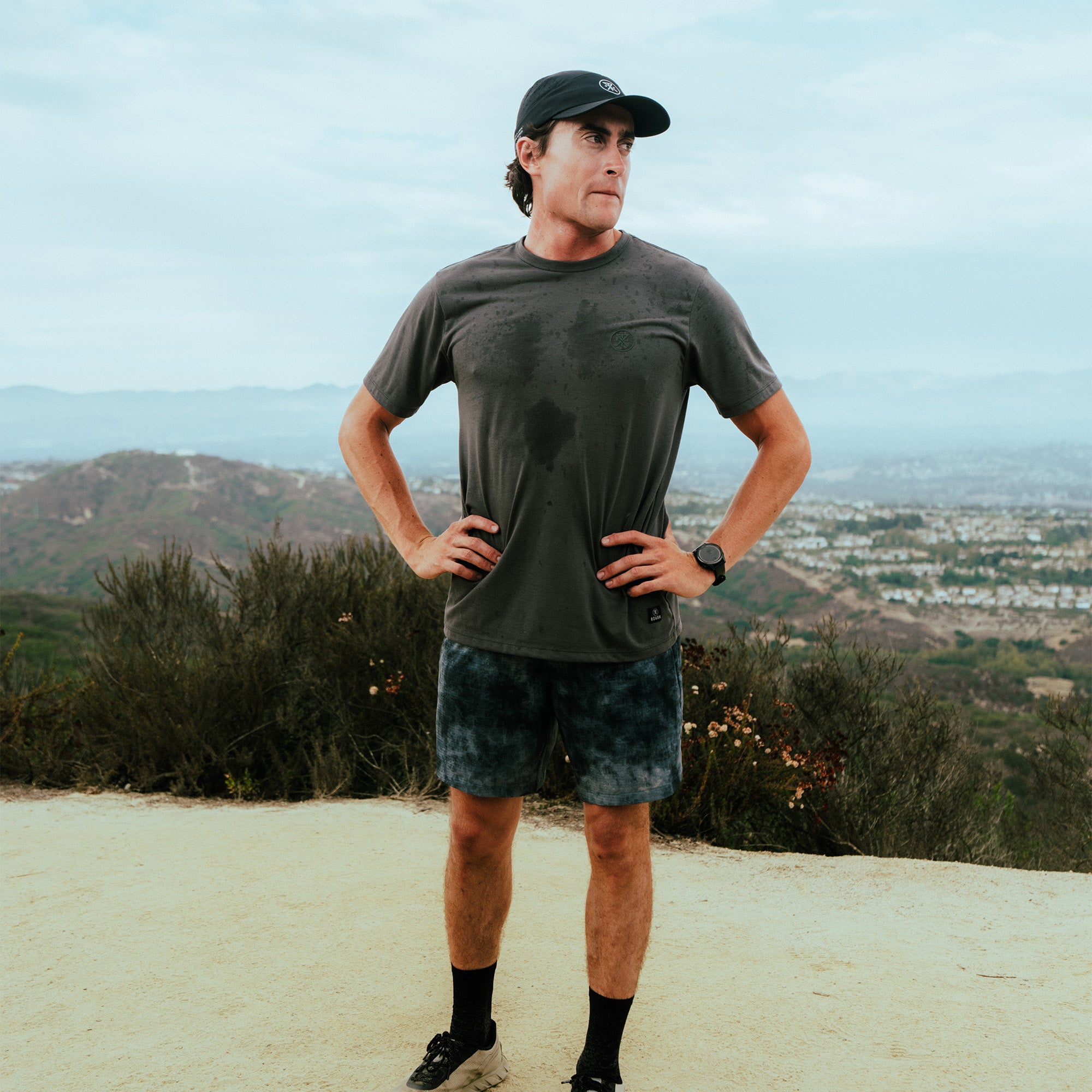 Man wearing charcoal short-sleeve t-shirt and denim shorts standing on hilltop overlook with valley landscape behind him