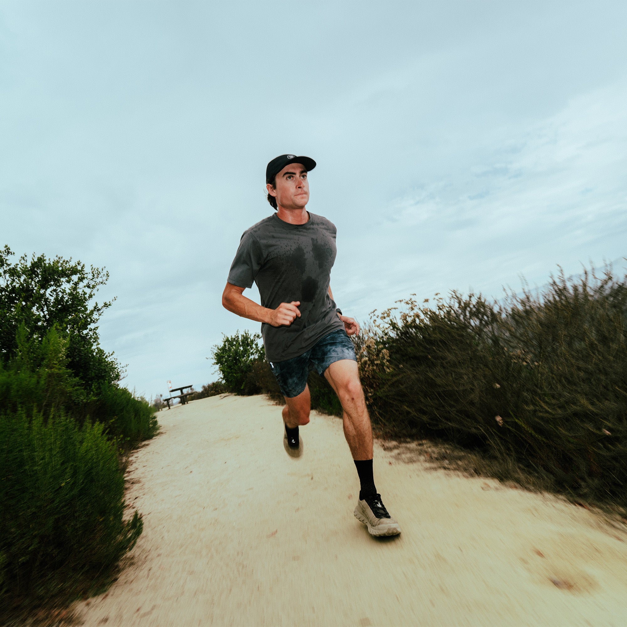 Man jogging on sandy trail wearing charcoal short sleeve tee shirt and black baseball cap under blue sky