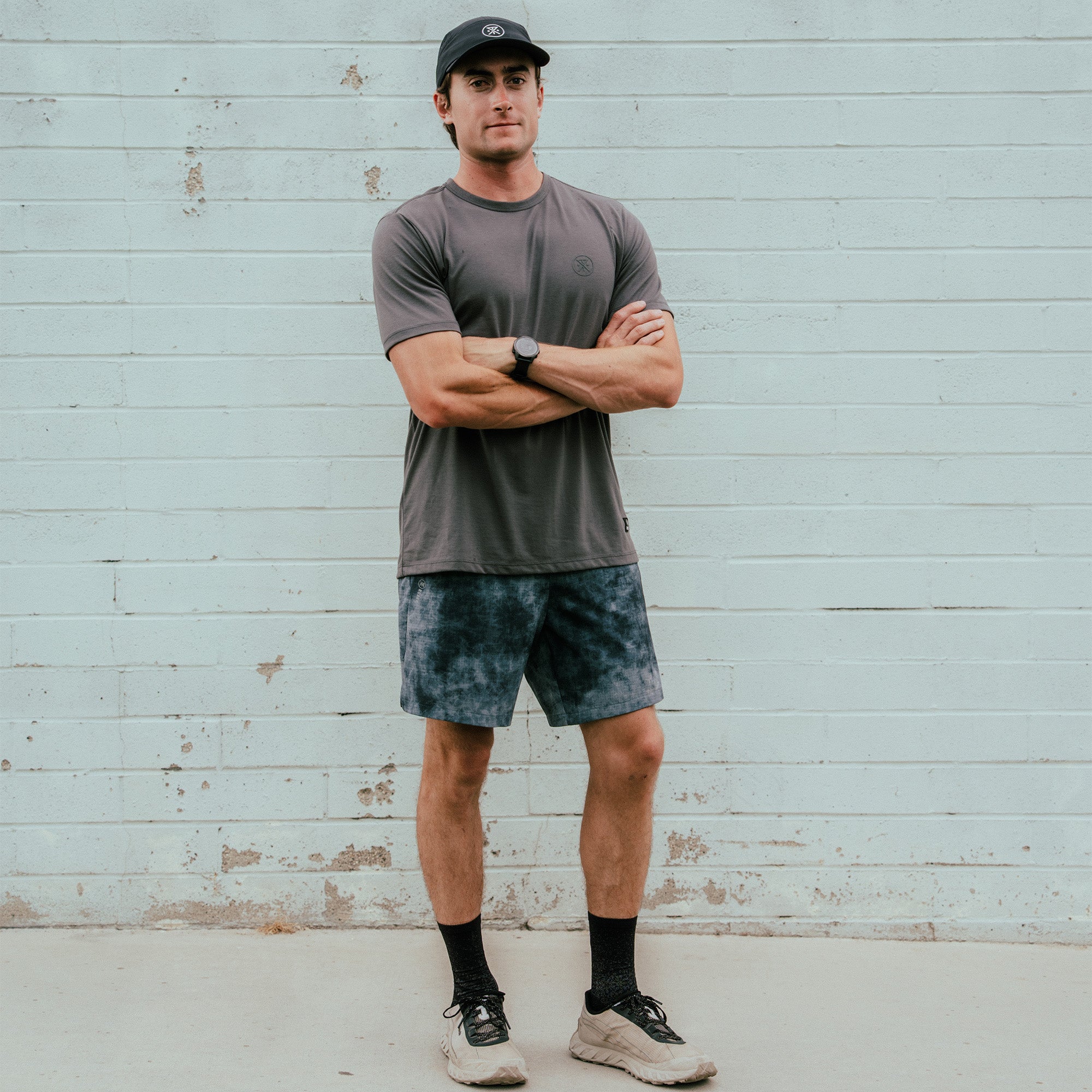 Man wearing charcoal gray short-sleeve tee with navy tie-dye shorts, black socks, and white sneakers against weathered wall