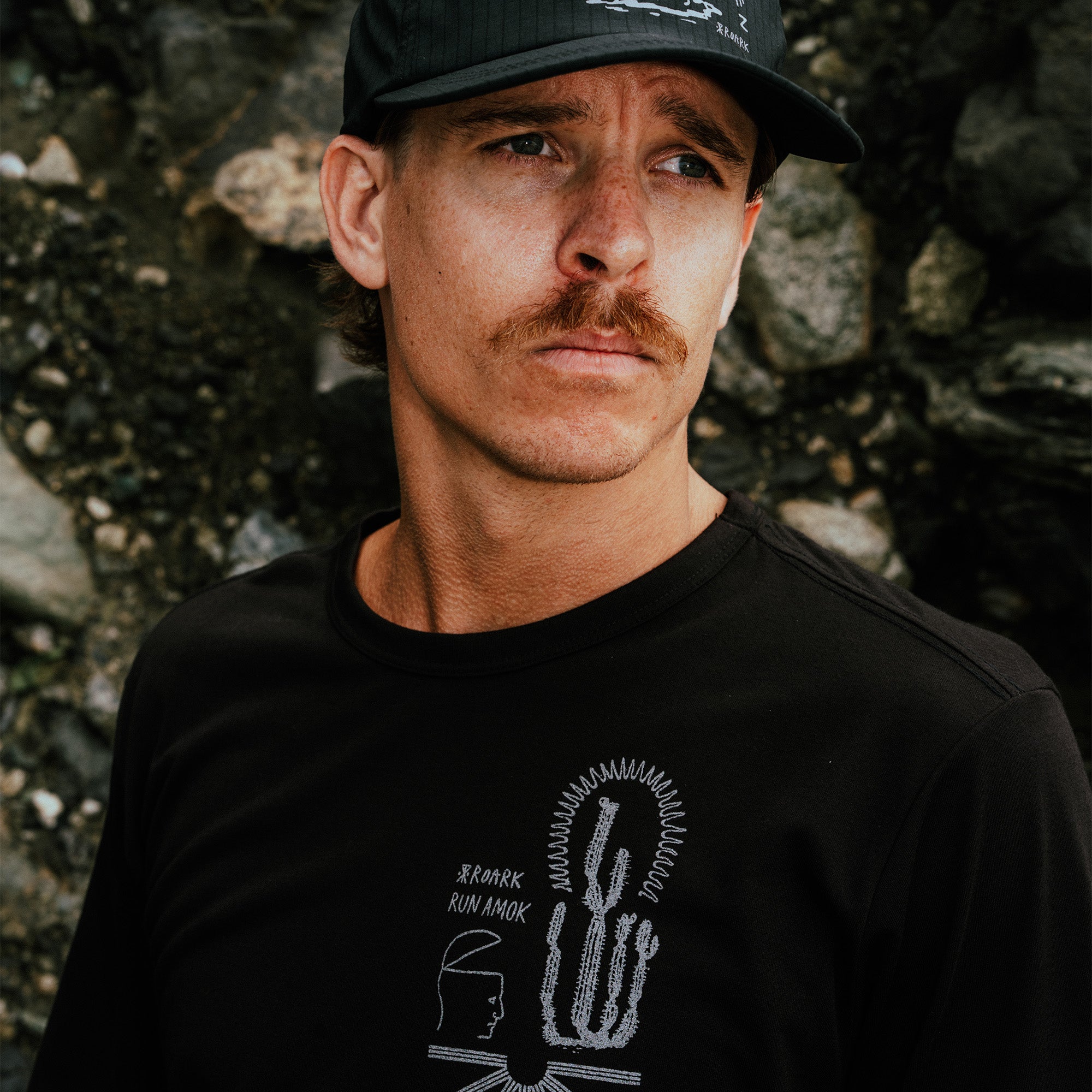 Black Mathis Active short sleeve tee with graphic print worn by man with black cap against textured stone wall