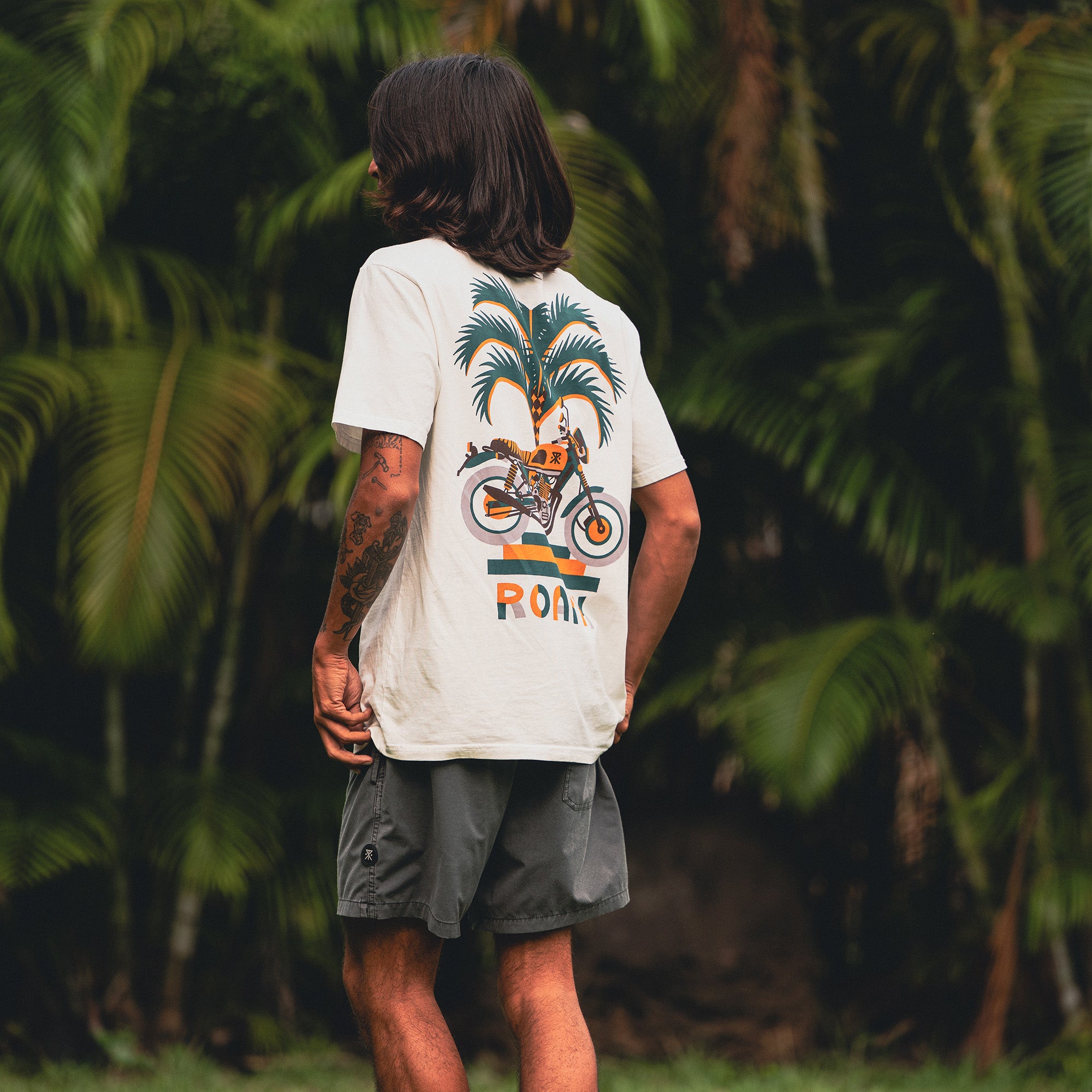 Person in a Moto Palm Premium Tee and boardshorts standing among palm trees, embodying Roark's spirit of adventure and travel.