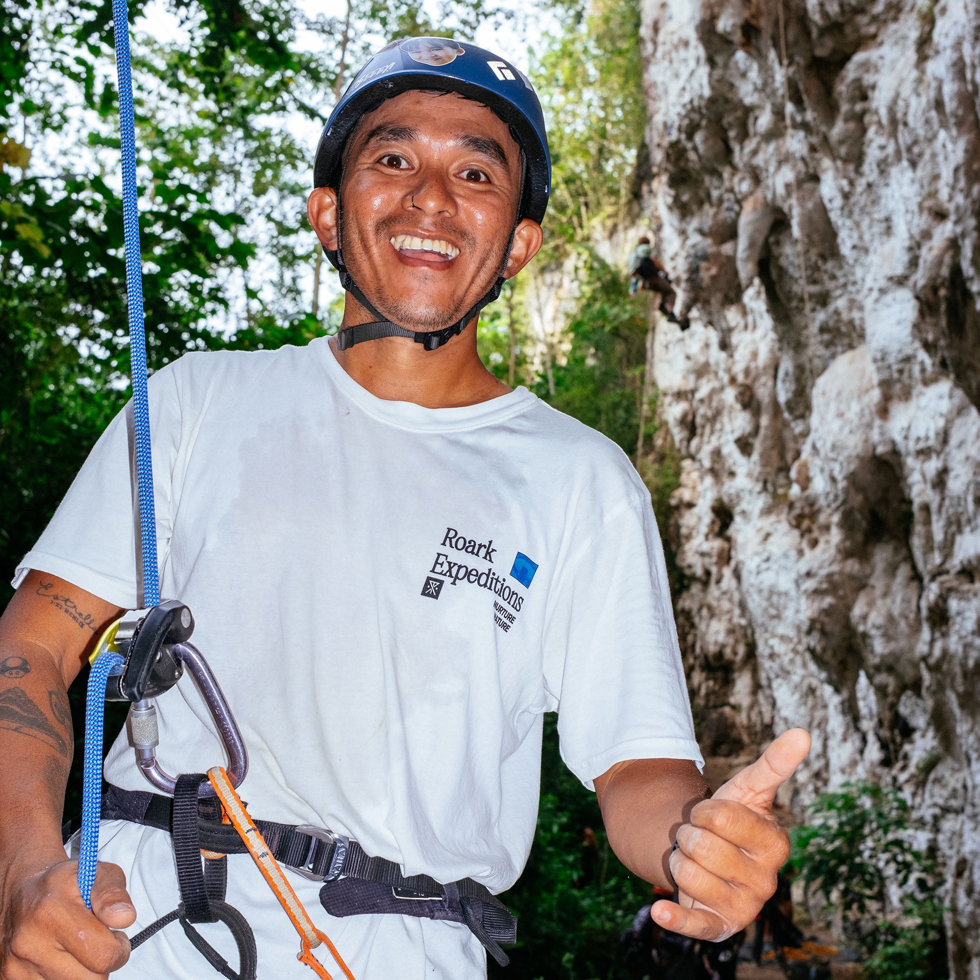 Rock Expeditions white t-shirt worn by smiling man with climbing gear and blue helmet against stone cliff