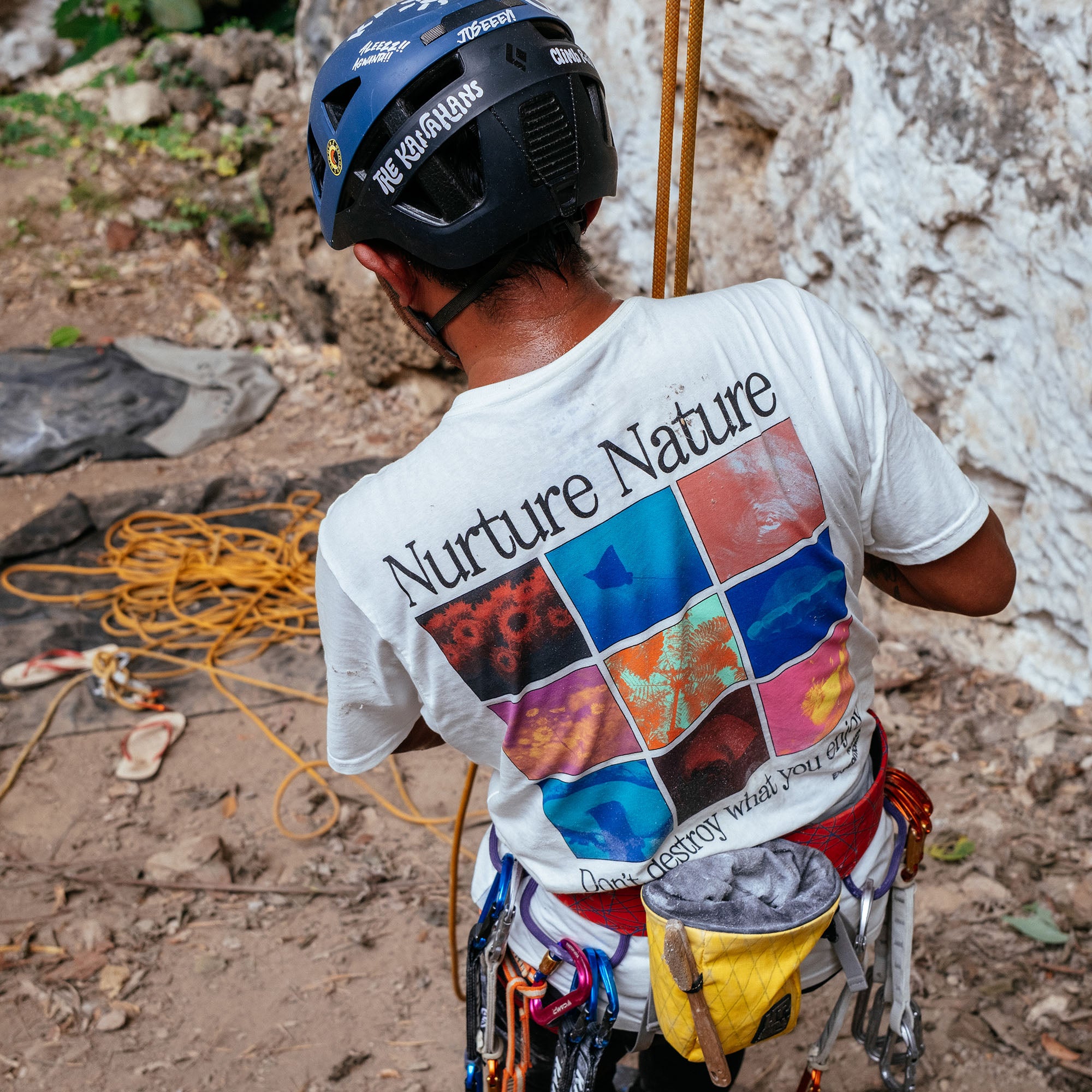 Climber wearing white Nurture Nature graphic tee with blue and black helmet, secured with safety gear at rocky cliff face