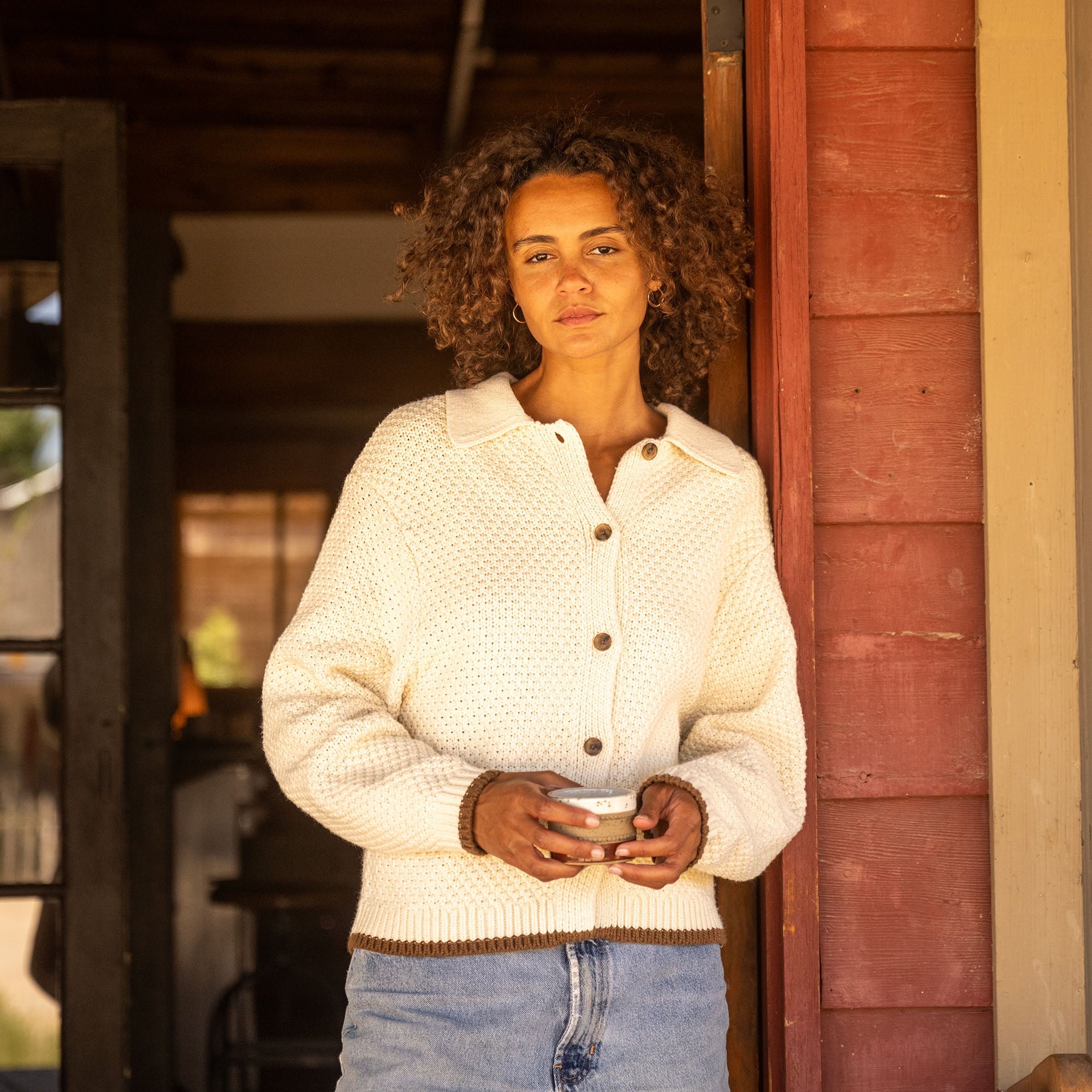 Woman wearing cream-colored knit cardigan with brown trim and button front over blue jeans, standing in doorway