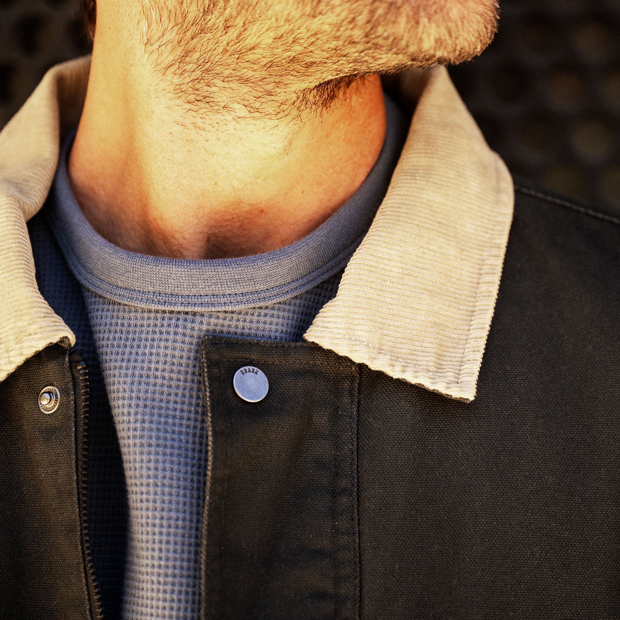 Black waxed canvas jacket with cream sherpa collar worn open over blue thermal shirt, detail view on male model